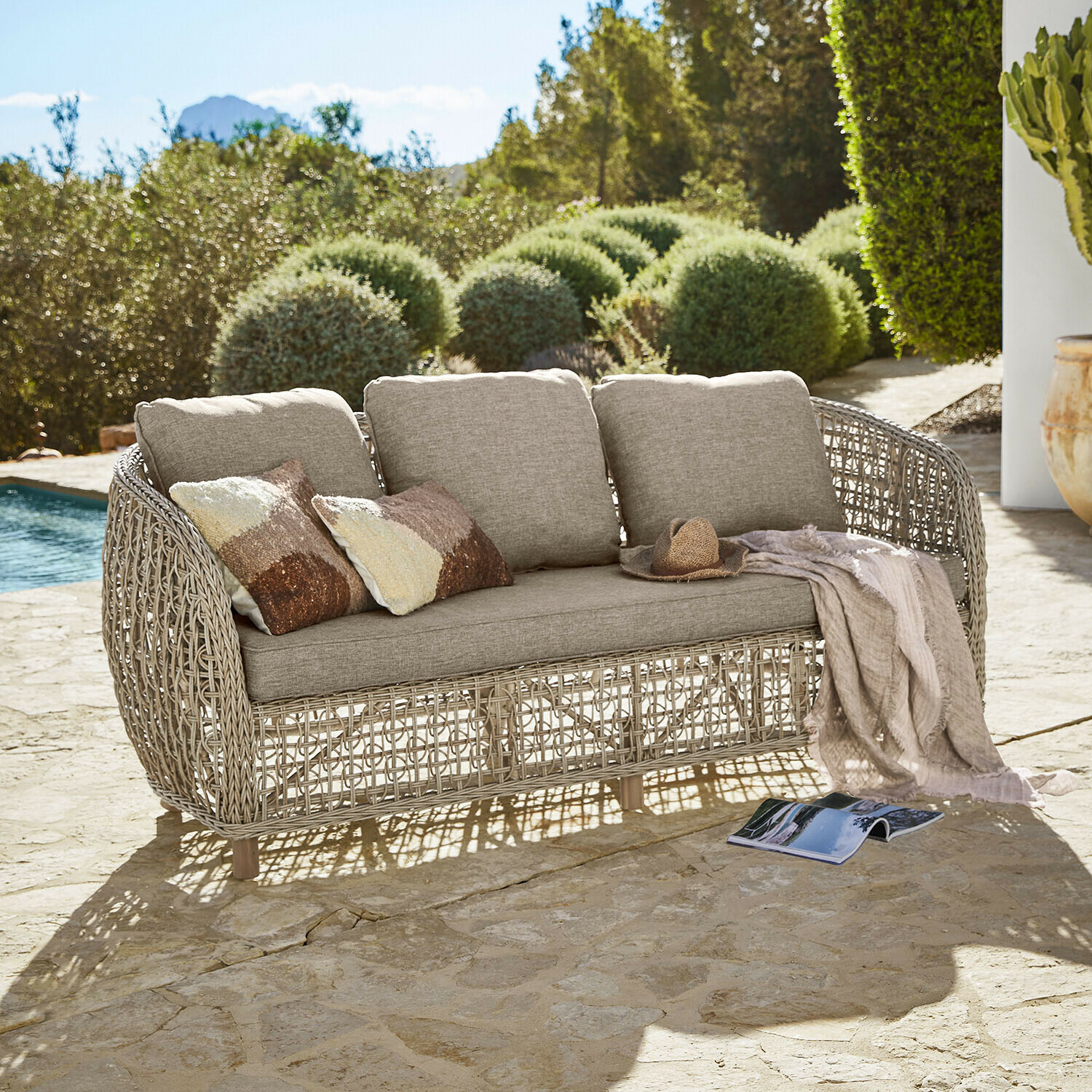 Buitensofa met beige kussens en gevlochten rotan frame op stenen terras bij zwembad
