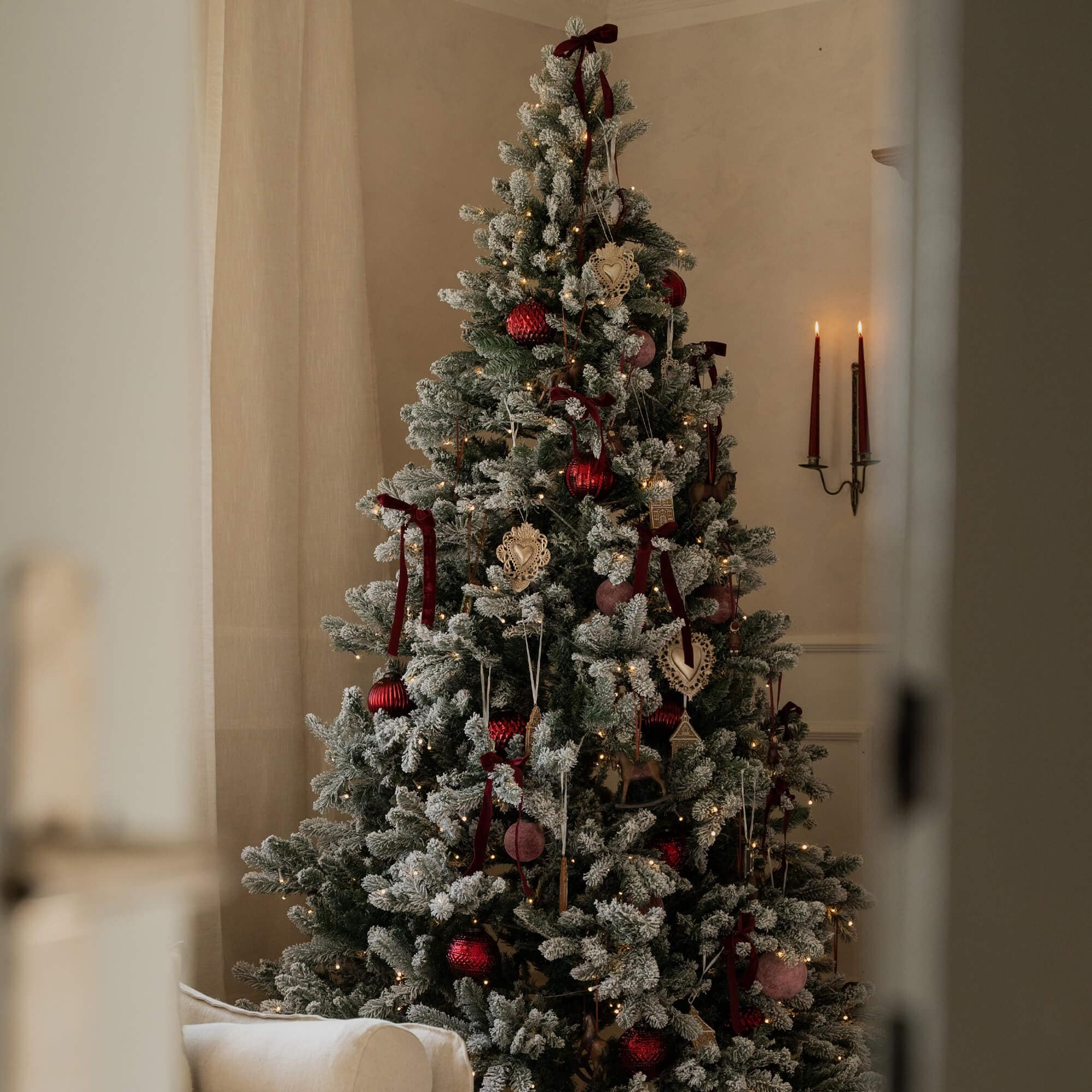 Besneeuwde kerstboom met rode en beige versieringen en warme verlichting in een gezellige kamer