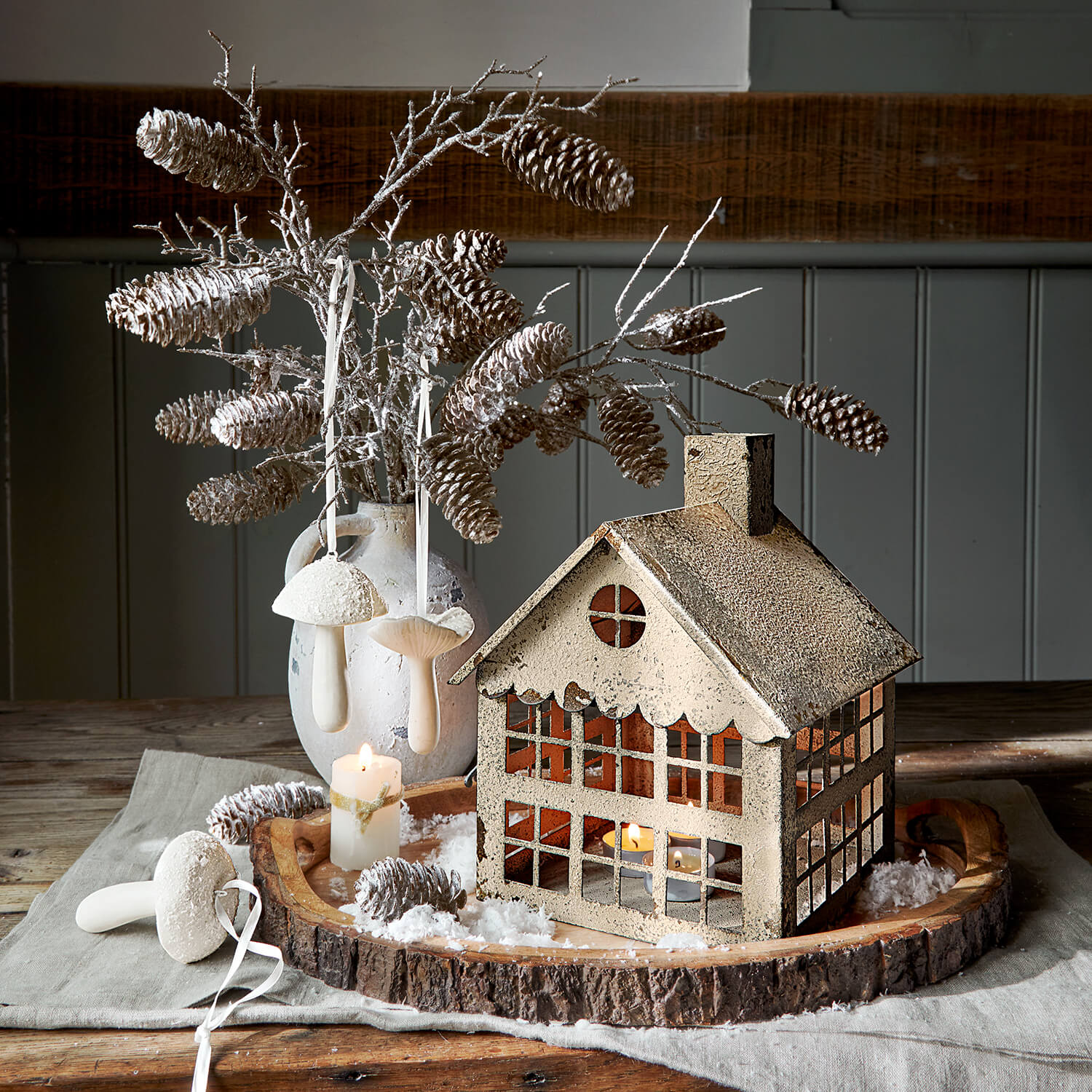 Beige metalen huisje als windlicht op houten dienblad met dennenappels en brandende kaars