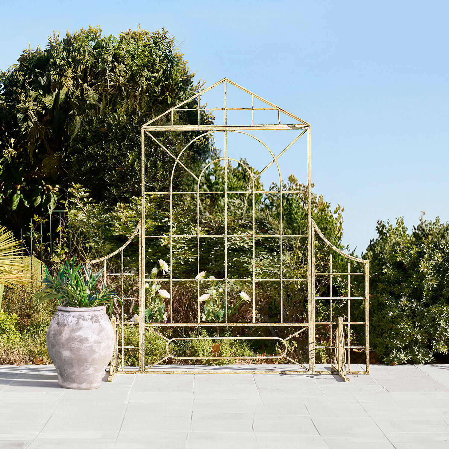Gouden metalen tuinrek in huisvorm op terras met pot en groen op de achtergrond