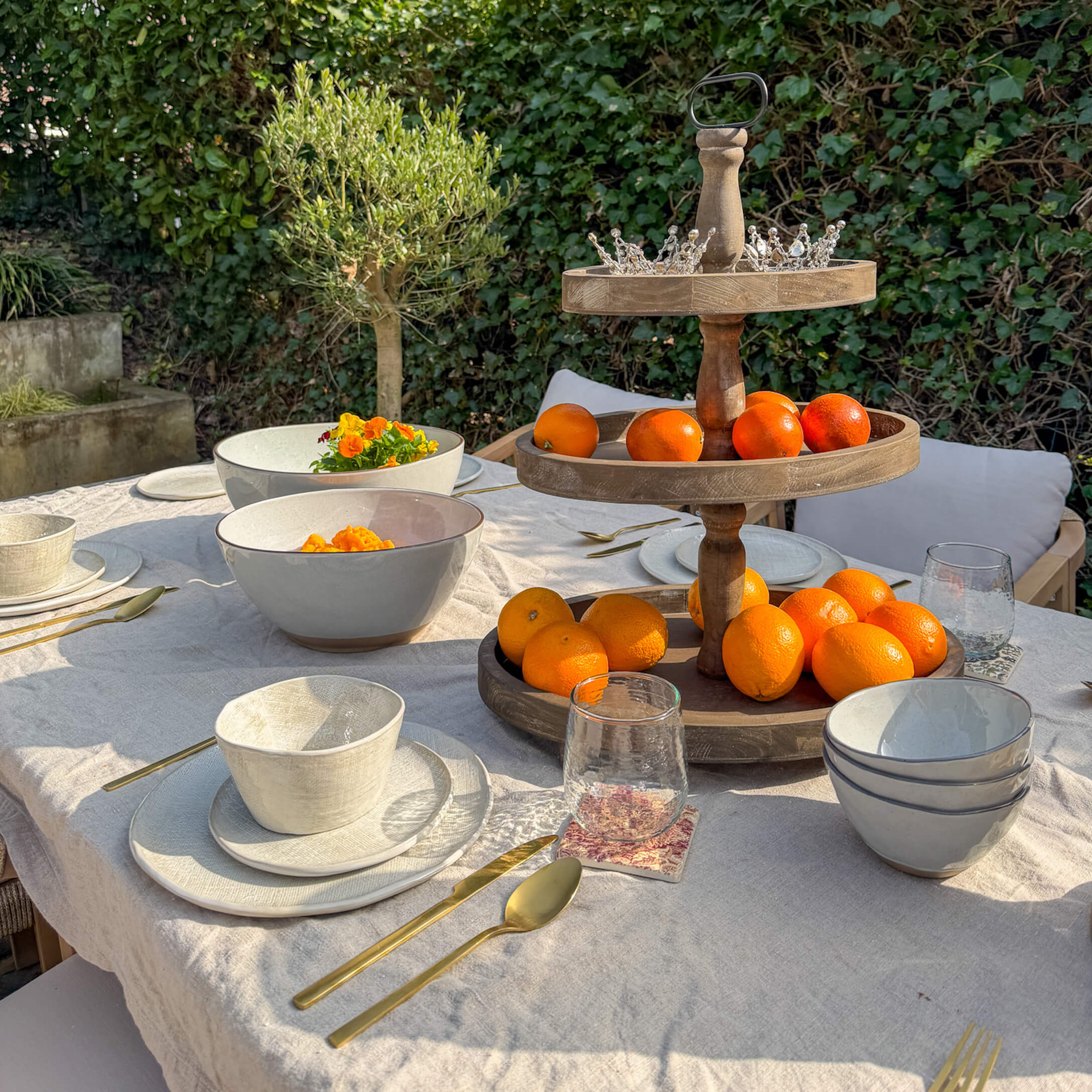 Tuin tafel met wit keramisch servies, gouden bestek en houten fruitstandaard met sinaasappels