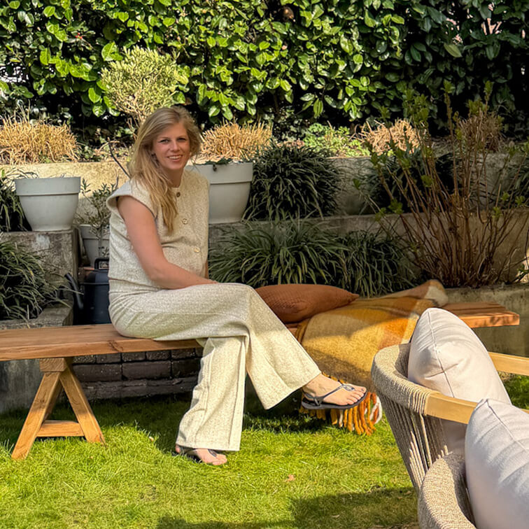 Vrouw in lichte outfit zittend op houten bank in tuin met groene planten en kussens