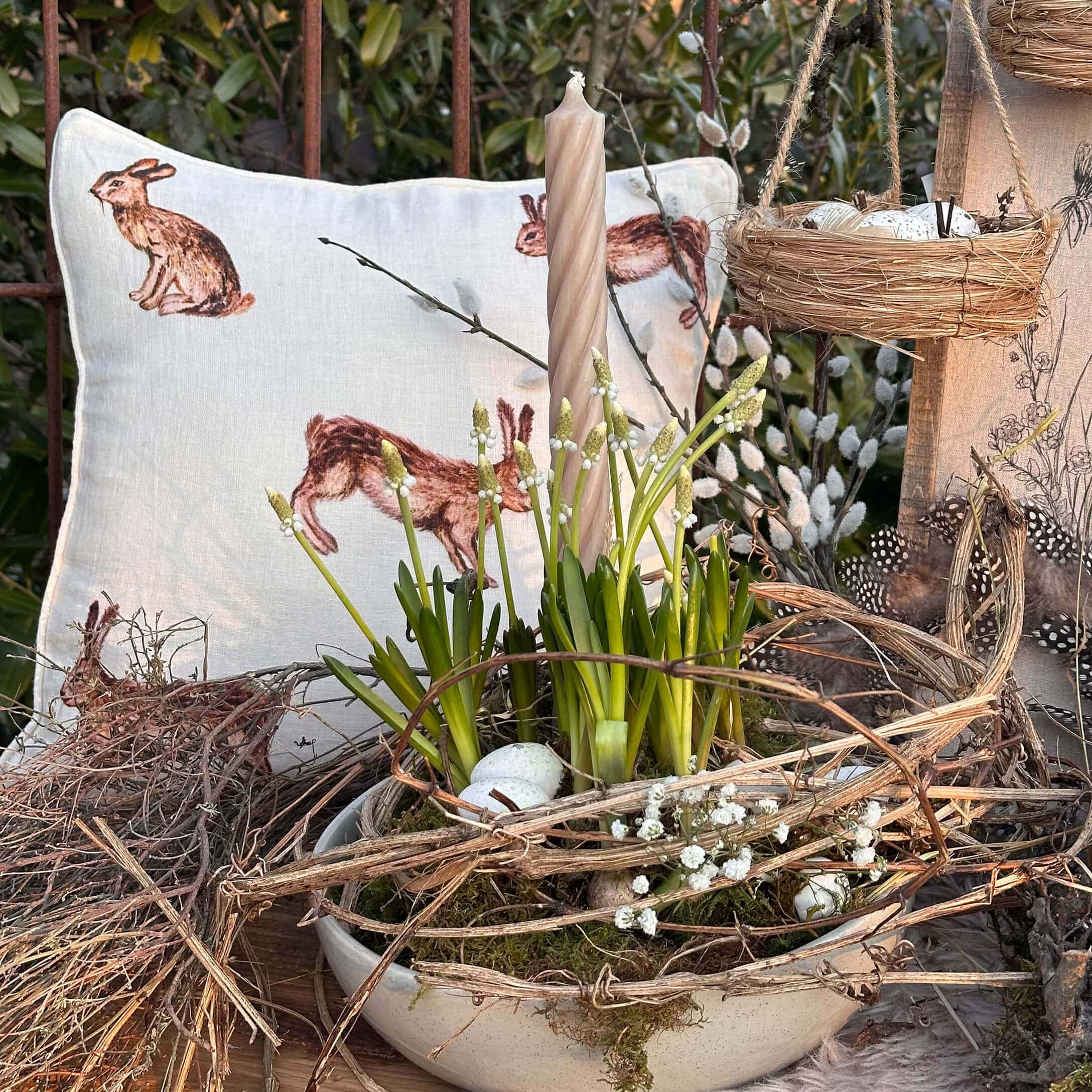 Natuurlijke paasdecoratie met witte kaars, groene bloemen en kussen met konijnenmotief op houten tafel