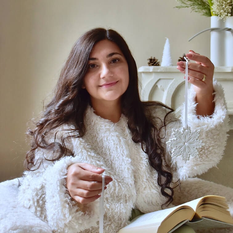 Vrouw in witte, pluizige trui houdt witte kerstversiering in sneeuwvlokvorm vast met open boek op schoot
