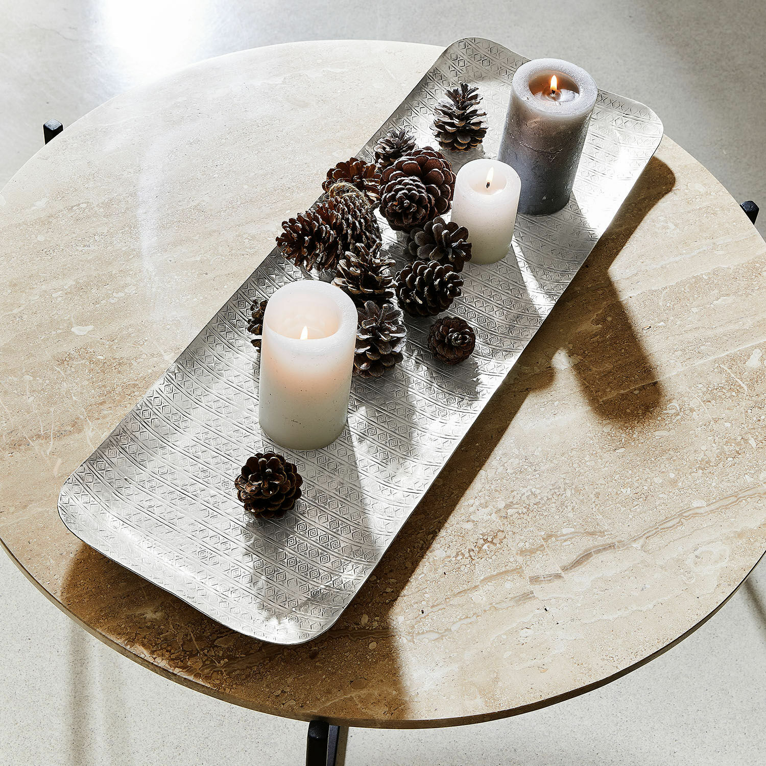 Zilveren metalen dienblad met geometrisch patroon, witte kaarsen en dennenappels op marmeren tafel