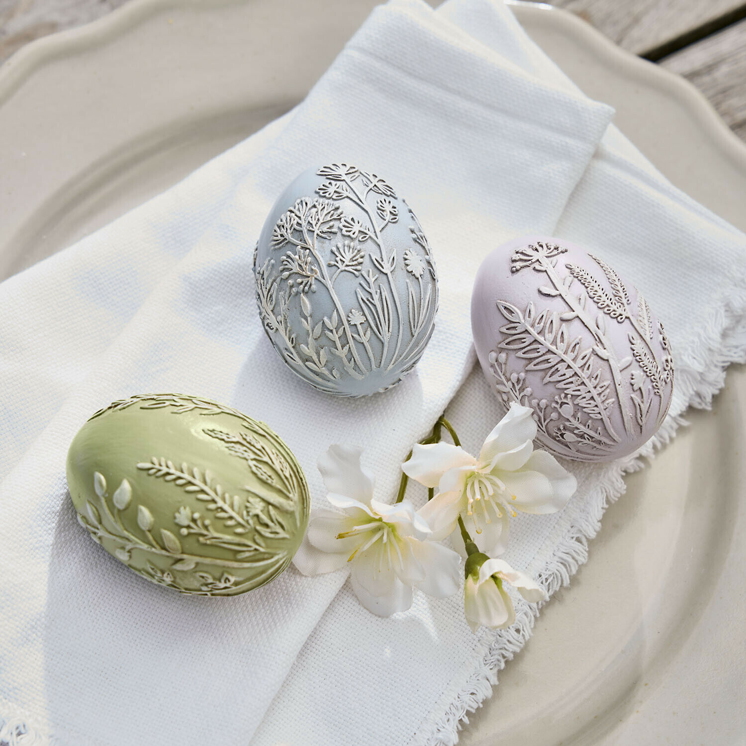 Drie pastelgekleurde decoratieve eieren met reliëf van planten op witte stof en bloemen op bord