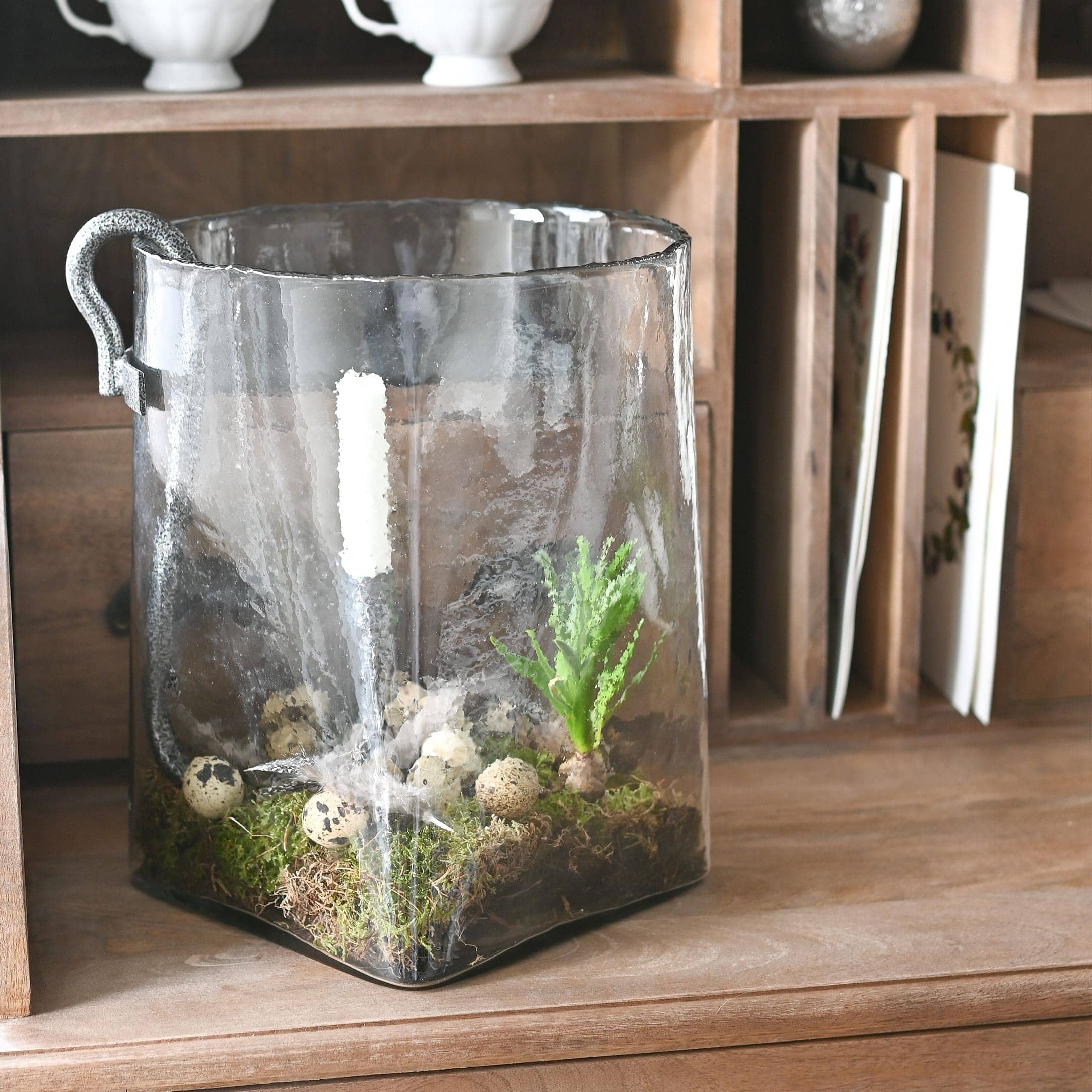 Transparante glazen container met handvat, versierd met mos en kwartel-eieren op een houten bureau