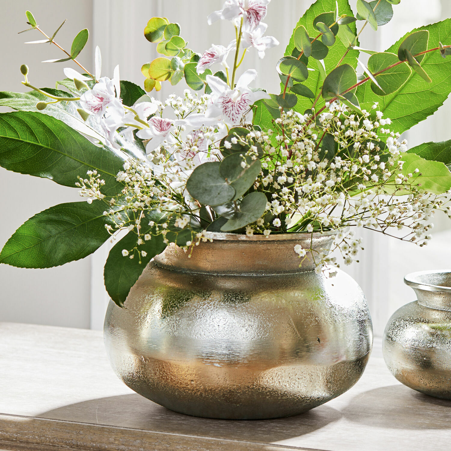 Vaas Hadrien Zilverkleurige metalen vaas met witte bloemen en groene bladeren op houten tafel, interieurdecoratie