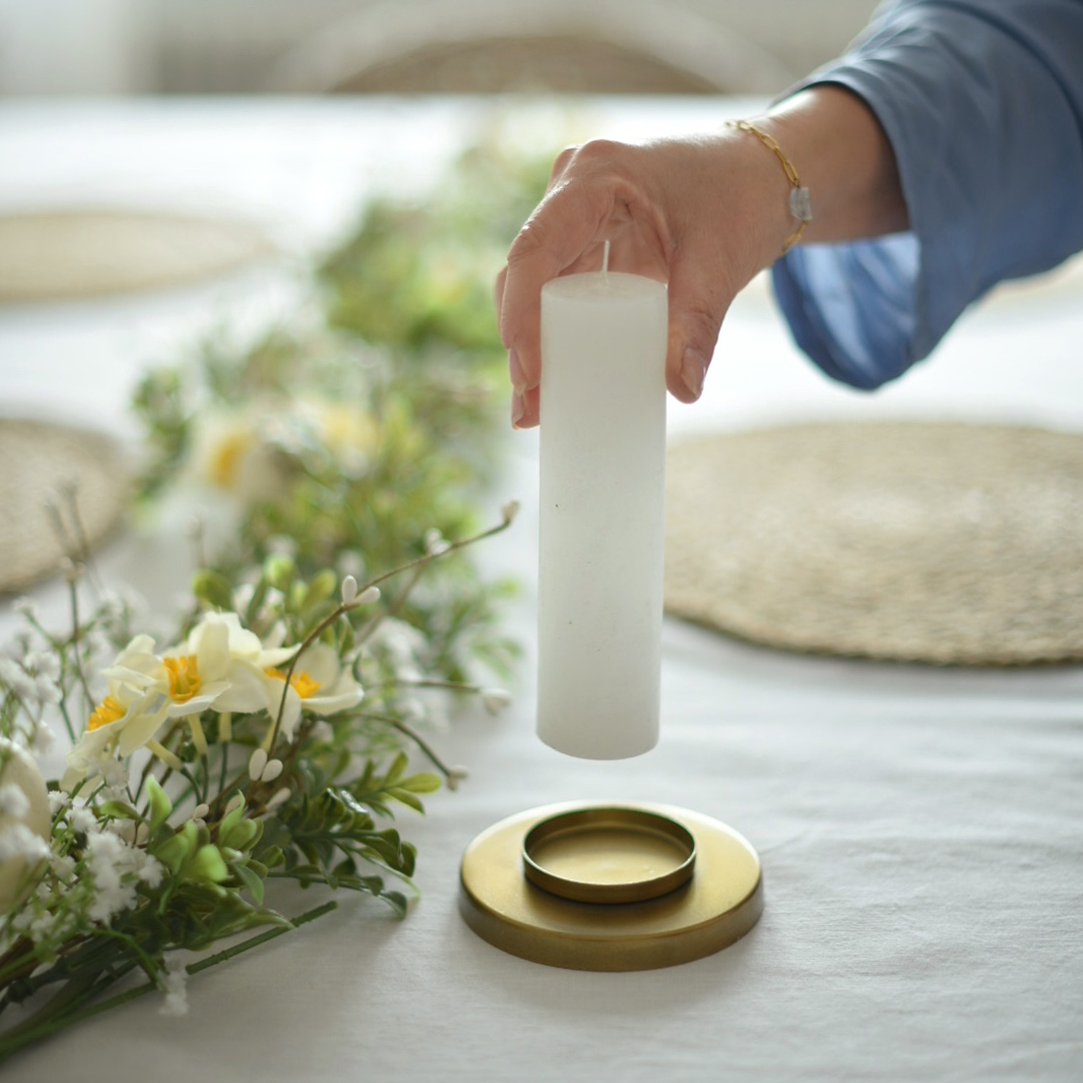 Witte stompkaars boven gouden kandelaar gehouden met bloemendecoratie op een wit tafelkleed