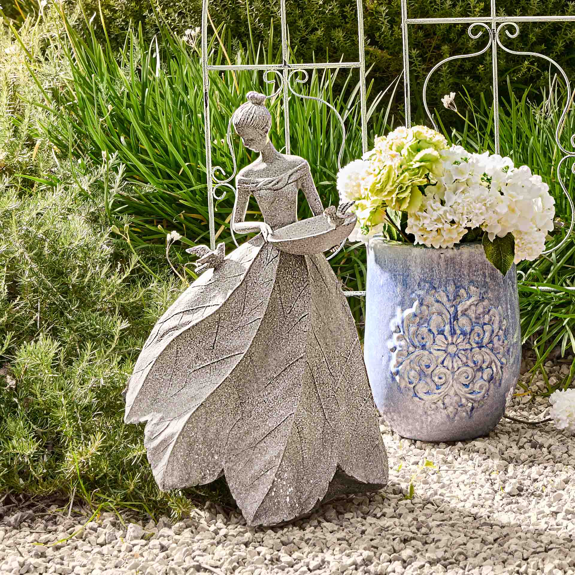 Decoratief vrouwenbeeld van grijs bladkeramiek naast blauwe vaas met witte bloemen in tuin