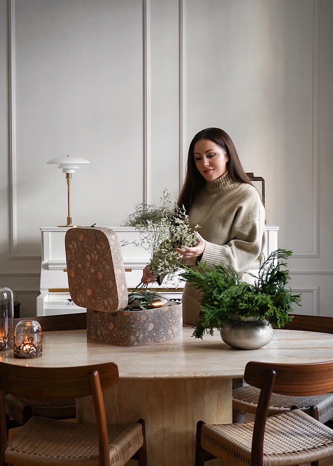 Vrouw in beige trui rangschikt bloemen aan marmeren tafel met decoratieve doos en groene planten