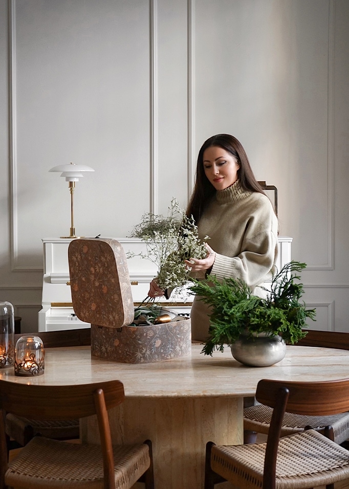 Vrouw in beige trui rangschikt bloemen aan marmeren tafel met decoratieve doos en groene planten