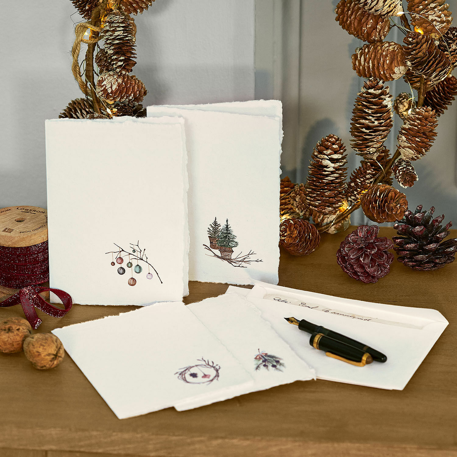 Wenskaarten set van 4 Velorine Set kerstkaarten van papier met dennenboom- en dennenappelmotieven op houten tafel met vulpen en decoraties