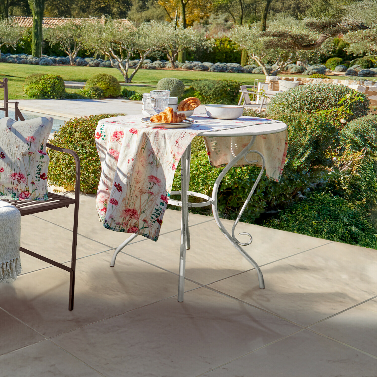 Tafel Aube Ronde witte metalen tuintafel met bloemen tafelkleed en gebak op terras met tuinzicht