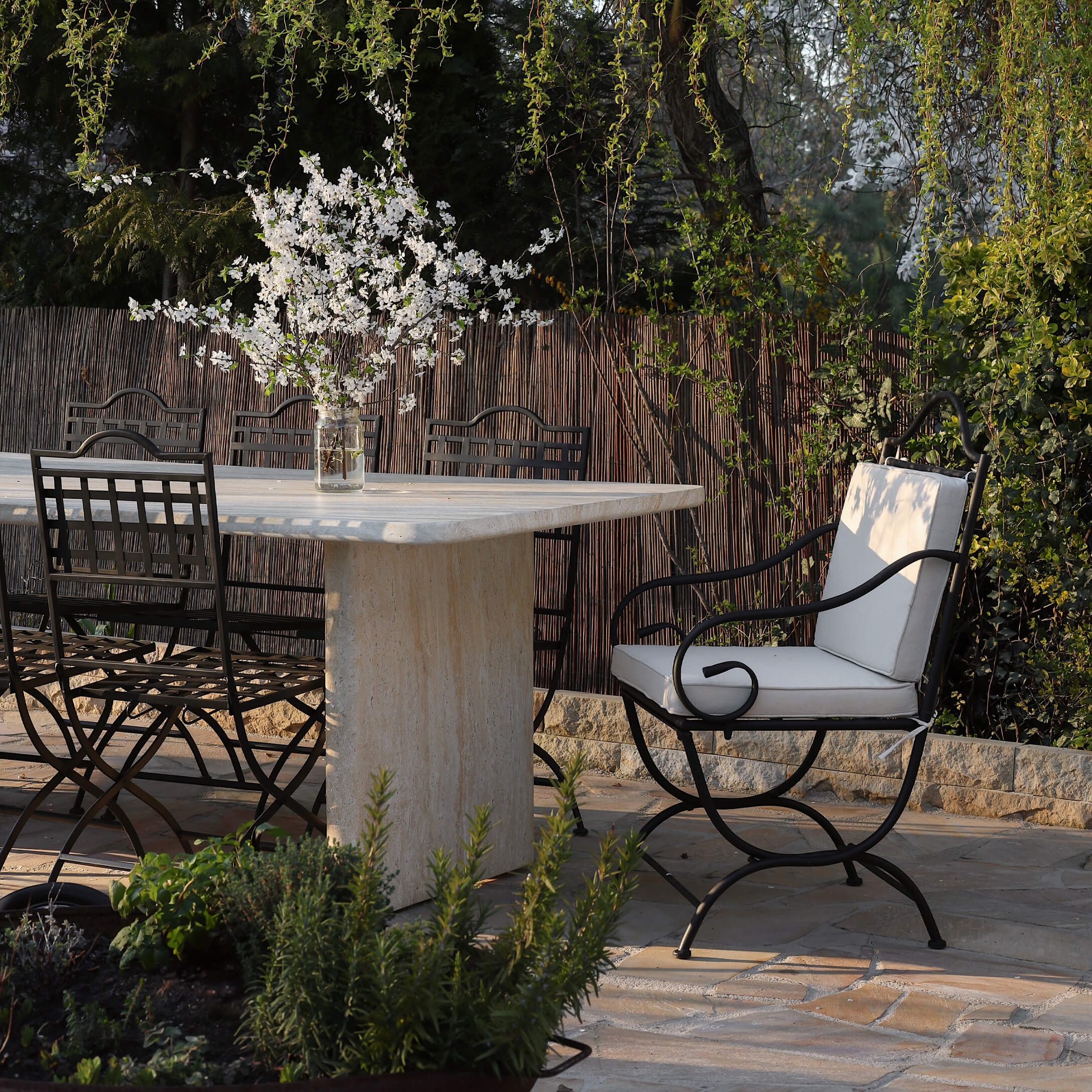 Tuin met stenen tafel en metalen stoelen met lichte kussens, bloemstuk op tafel