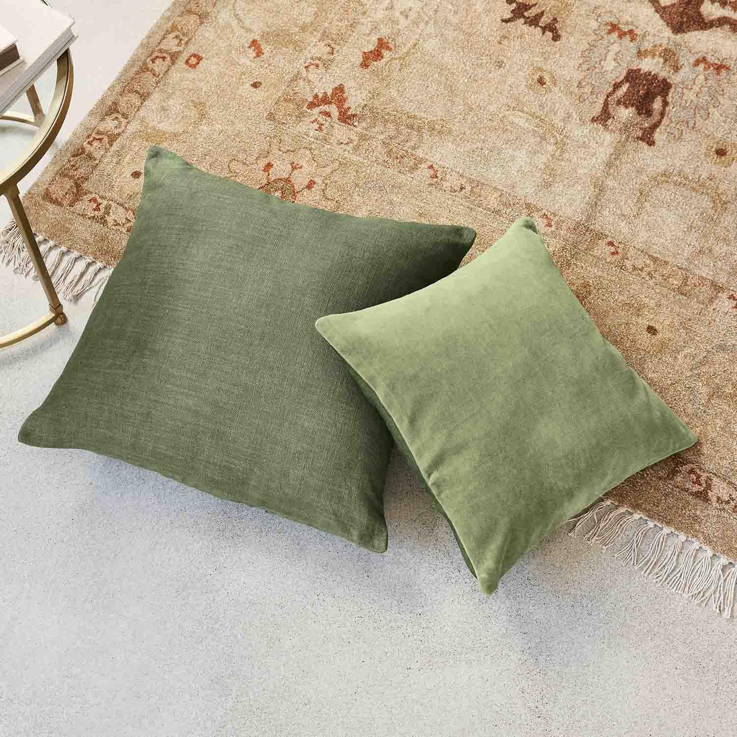 Twee groene stoffen kussens op een beige vloerkleed met franjes, stijlvolle woonaccessoires voor de woonkamer