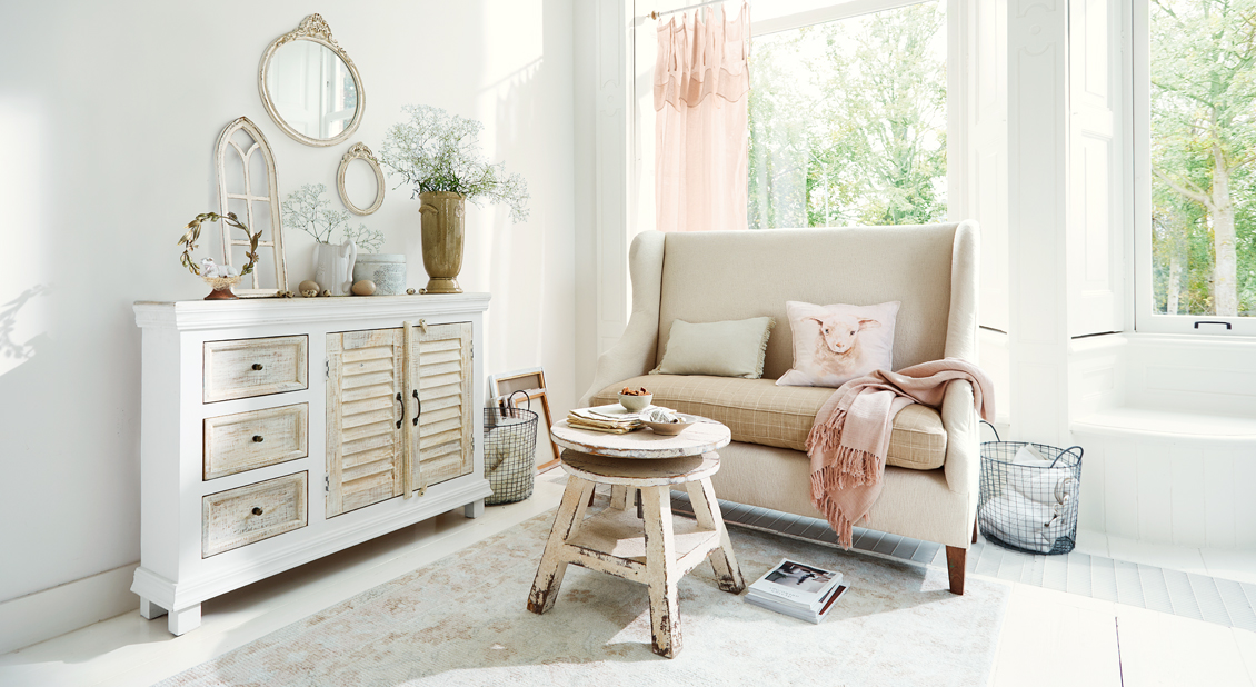 Gezellige shabby chic woonkamer met witte houten dressoir en beige bank met kussens en roze deken