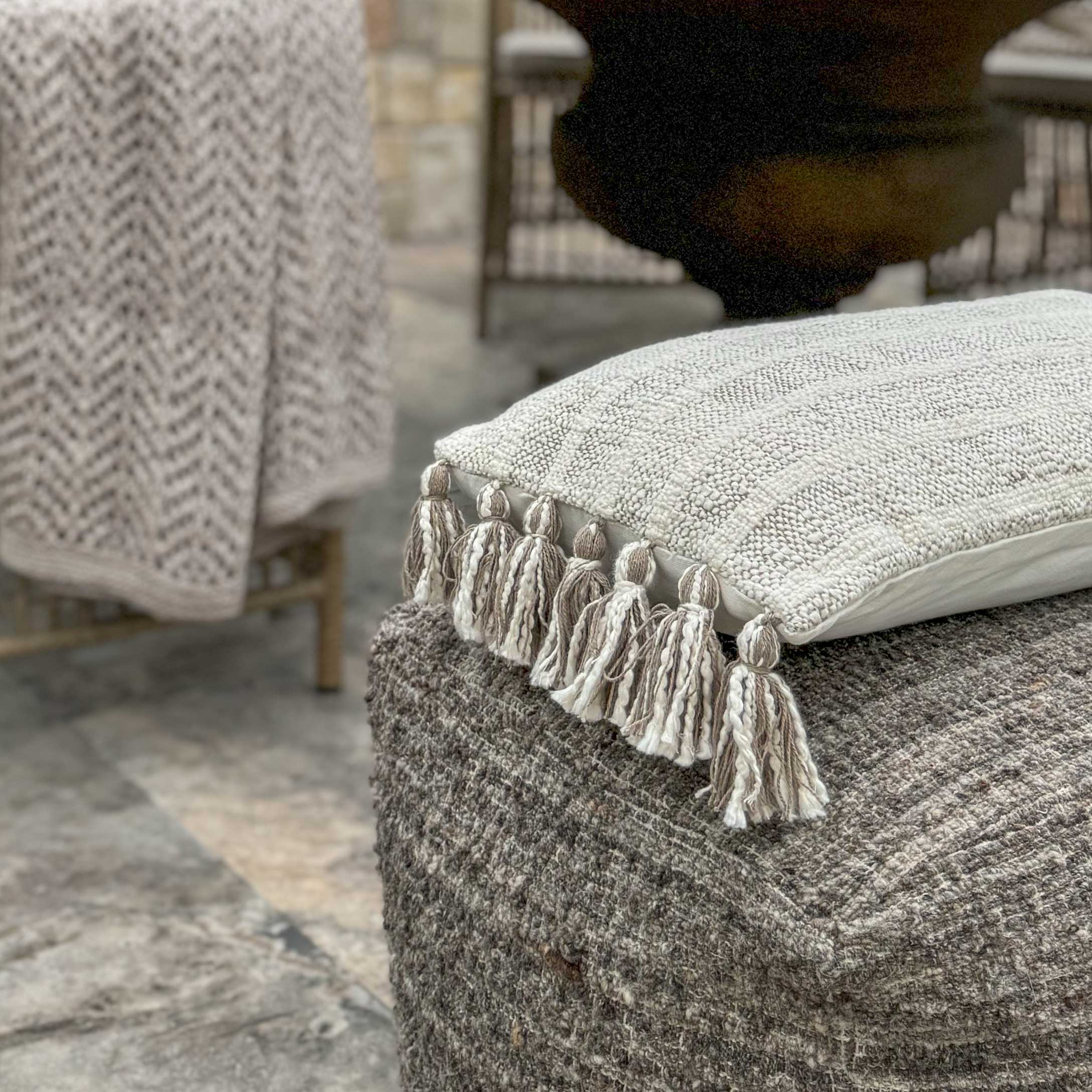 Beige decoratief kussen met kwastjes op een grijze, gestructureerde poef in boho stijl