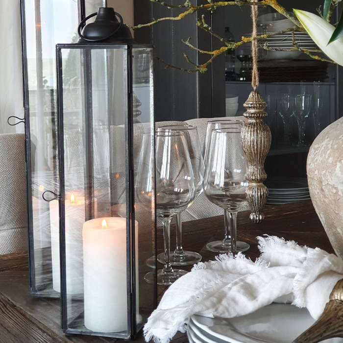 Glazen lantaarn met zwart metalen frame en witte kaars op houten tafel met glazen en witte doek