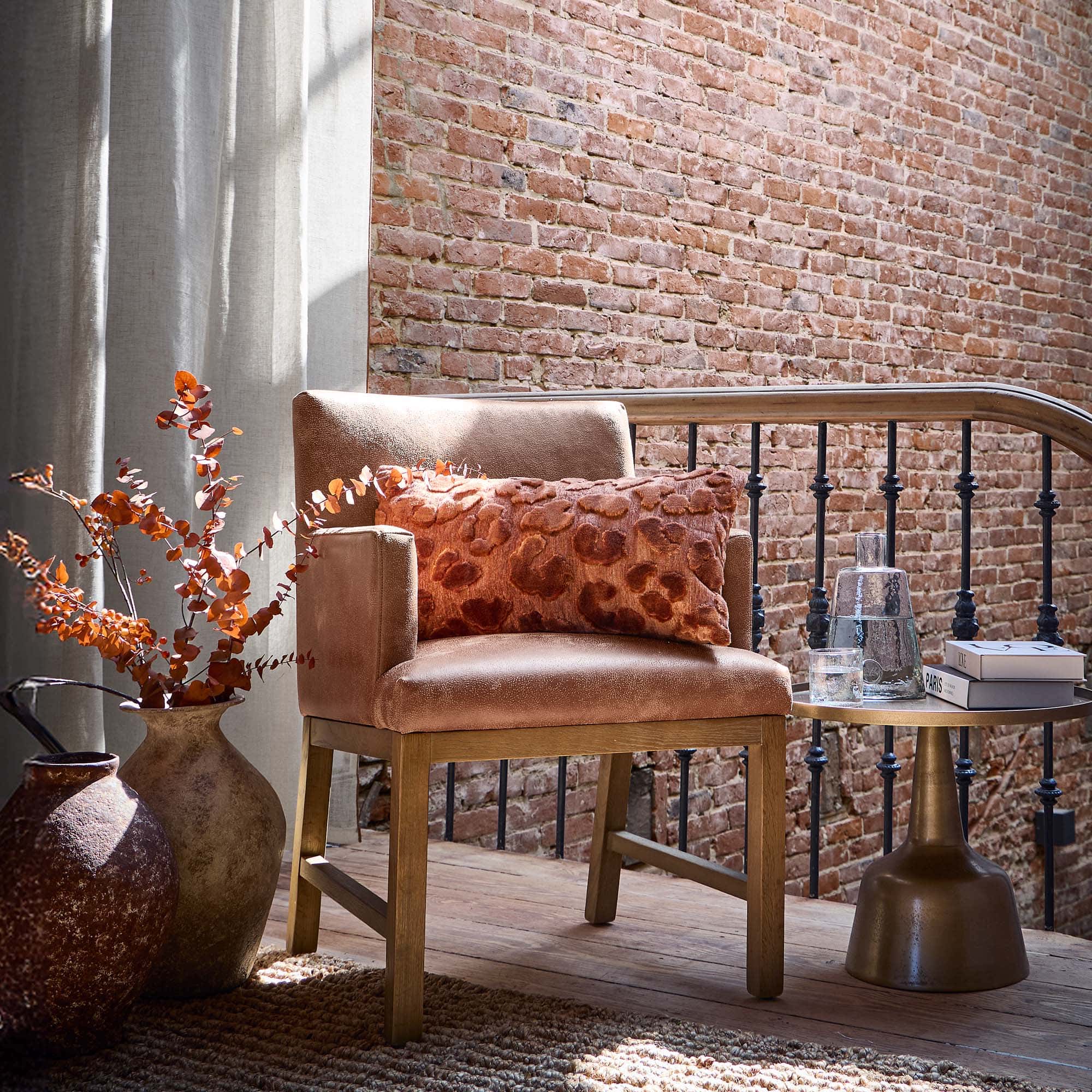 Beige fauteuil met houten onderstel en oranje kussen voor een bakstenen muur en gordijnen