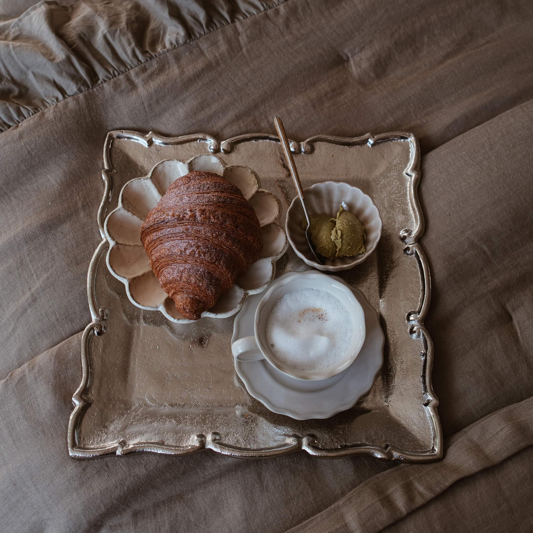 Dienblad met croissant, schuimkoffie en pasta op beige stof, slow living sfeer