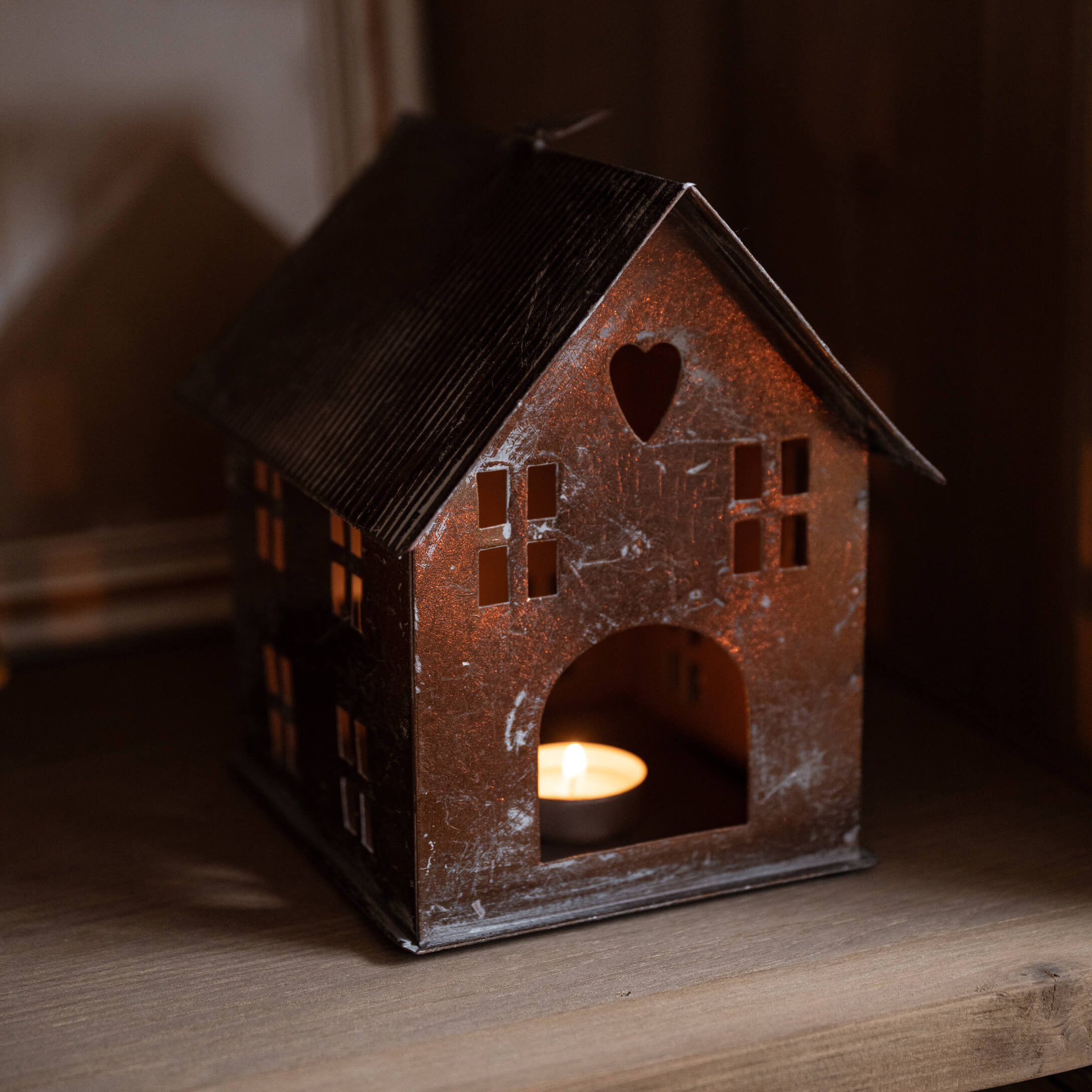 Metalen lantaarn in huisvorm met hartvormige uitsnede en brandende kaars binnen op houten plank