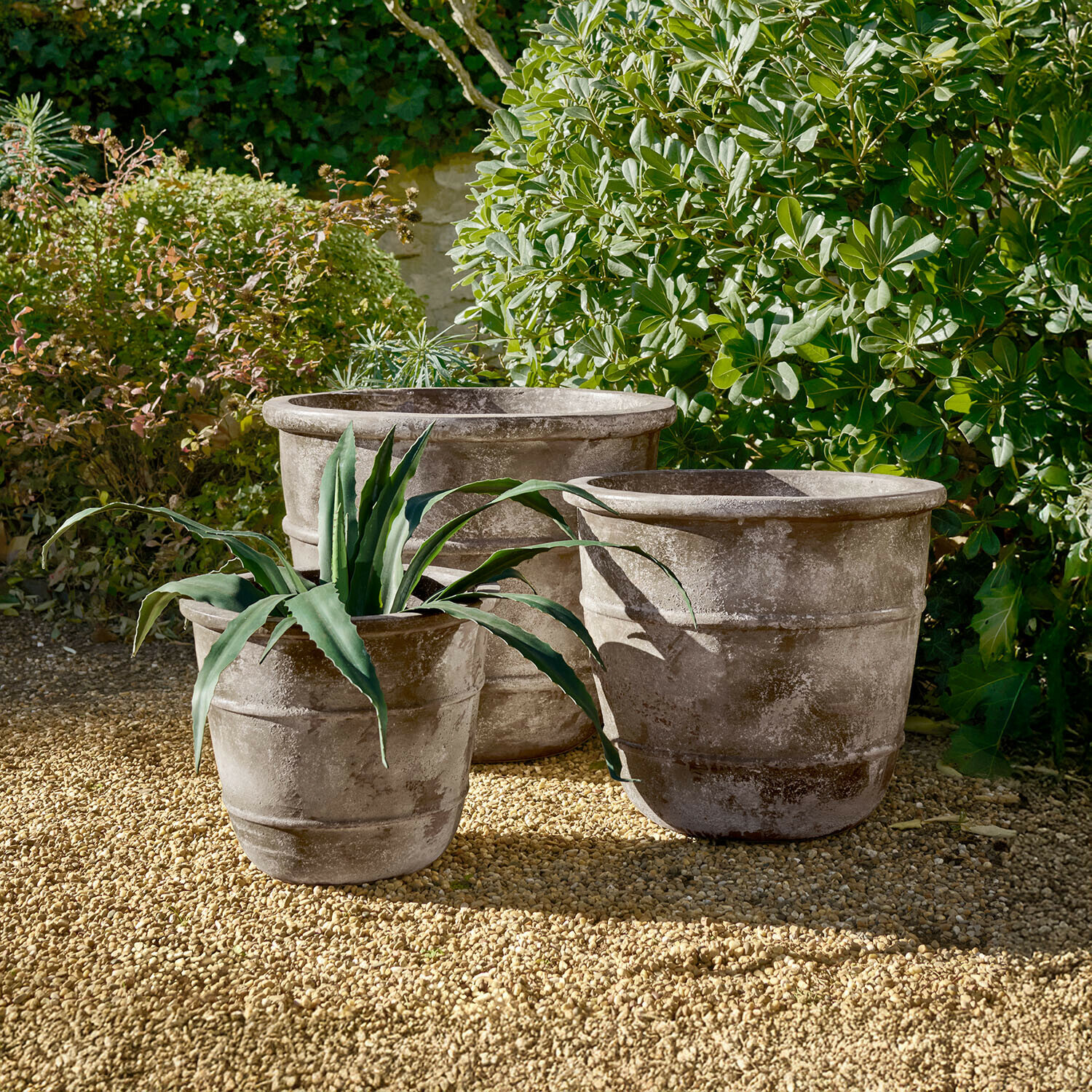 Drie grote grijze stenen potten op grind in een tuin met groene planten op de achtergrond
