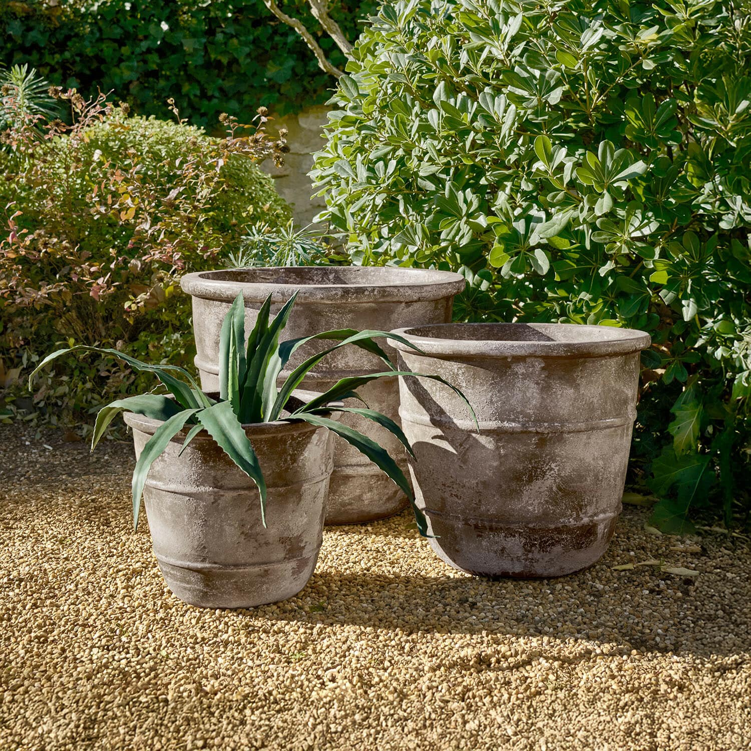 Sierpot set van 3 Arlivonne Drie grote grijze stenen potten op grind in een tuin met groene planten op de achtergrond