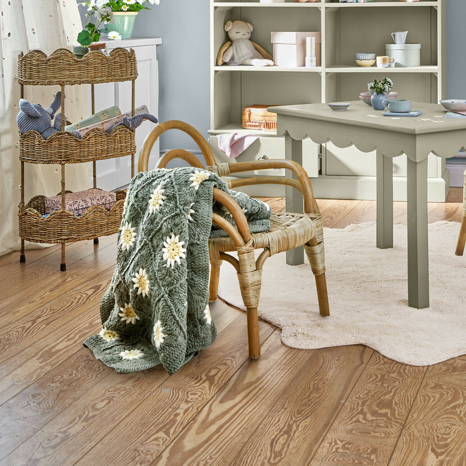 Grijze gebreide deken met witte bloemen op rotan stoel in lichte kinderkamer met houten vloer