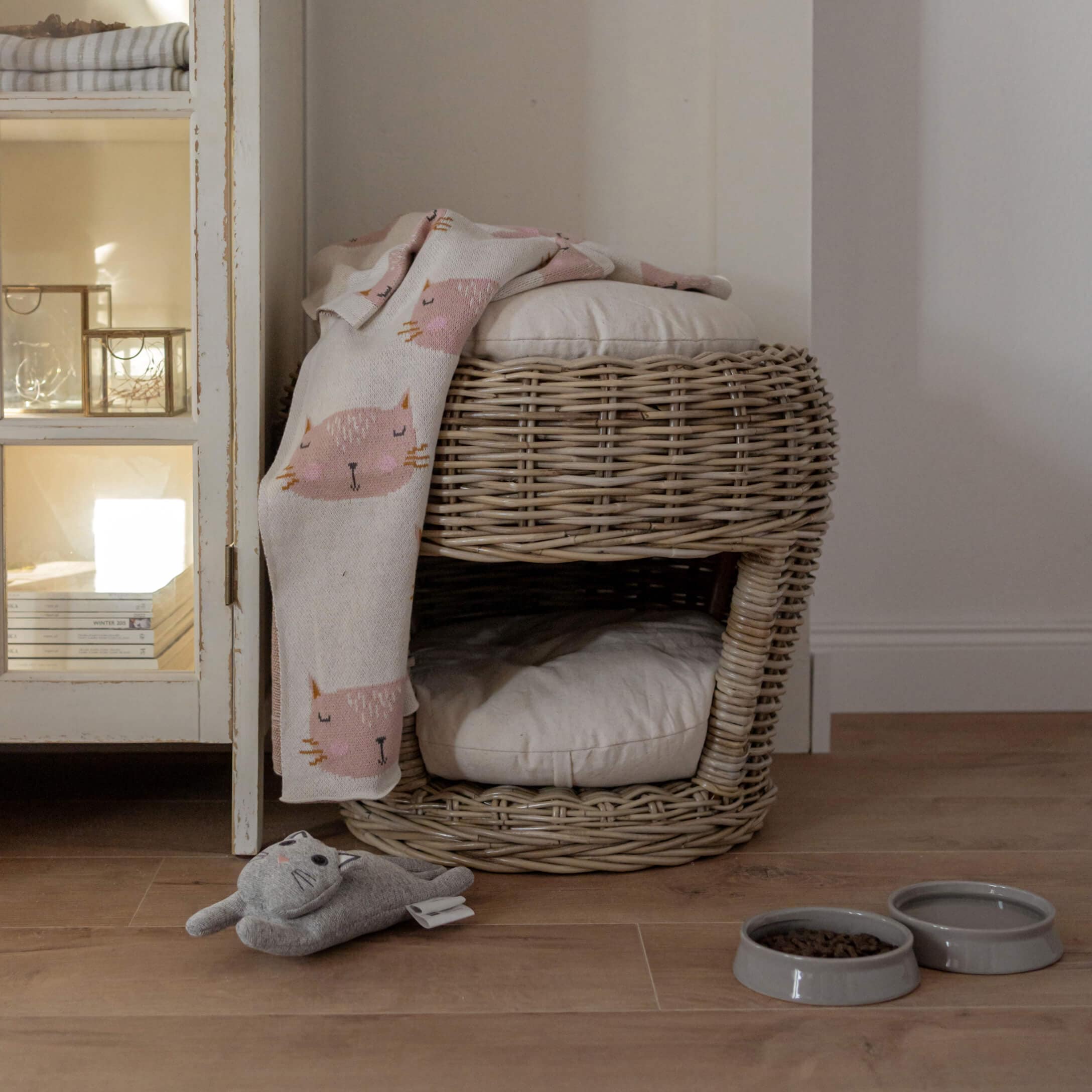 Tweedelig rotan kattenbed met lichte kussens, roze kattendeken, speelgoed en voerbakken