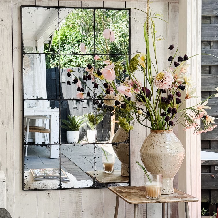 Wandspiegel met verdeelde panelen die bloemen in keramische vaas en koffie op rustieke houten tafel weerspiegelt