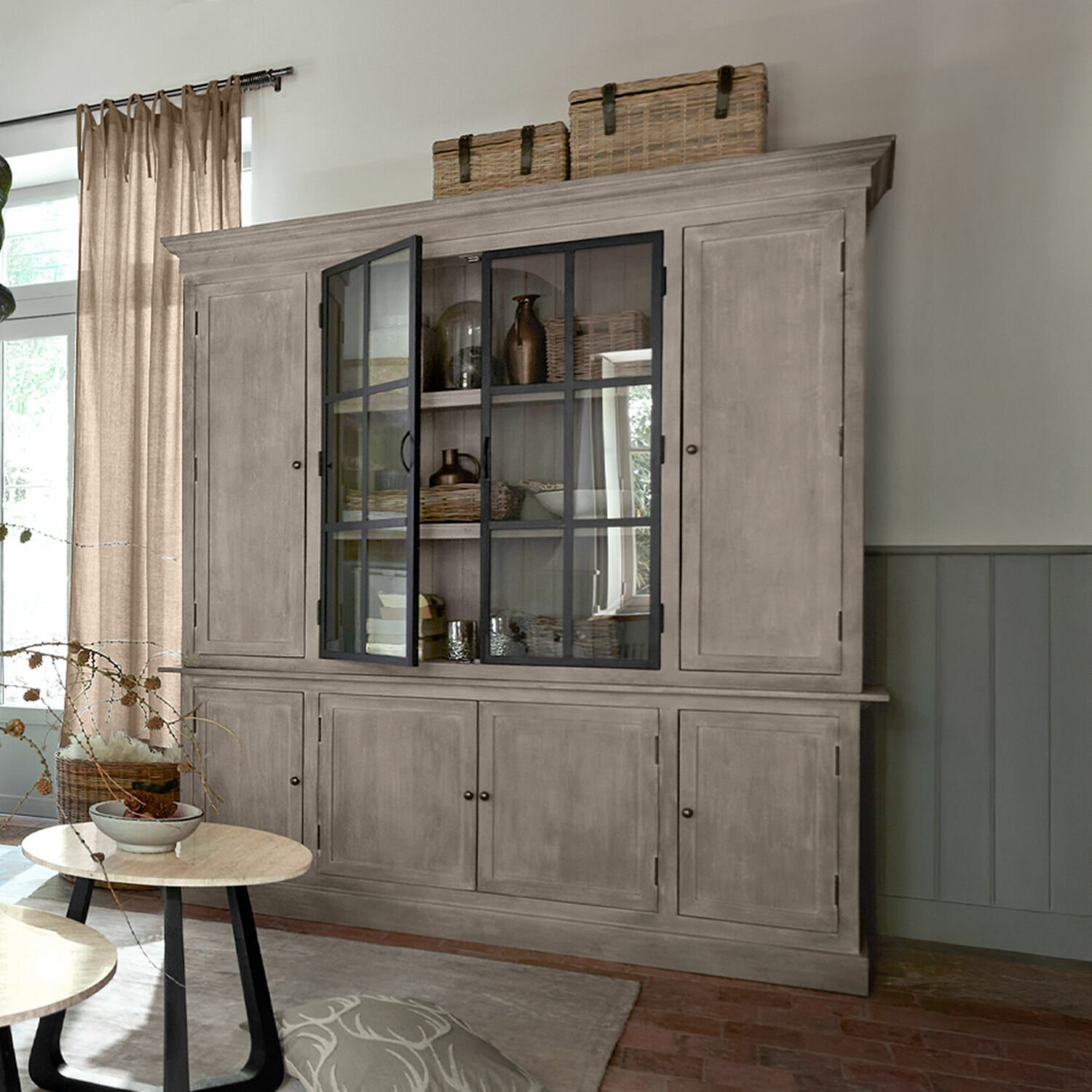 Grote grijze houten vitrinekast met glazen deuren en manden bovenop in woonkamer