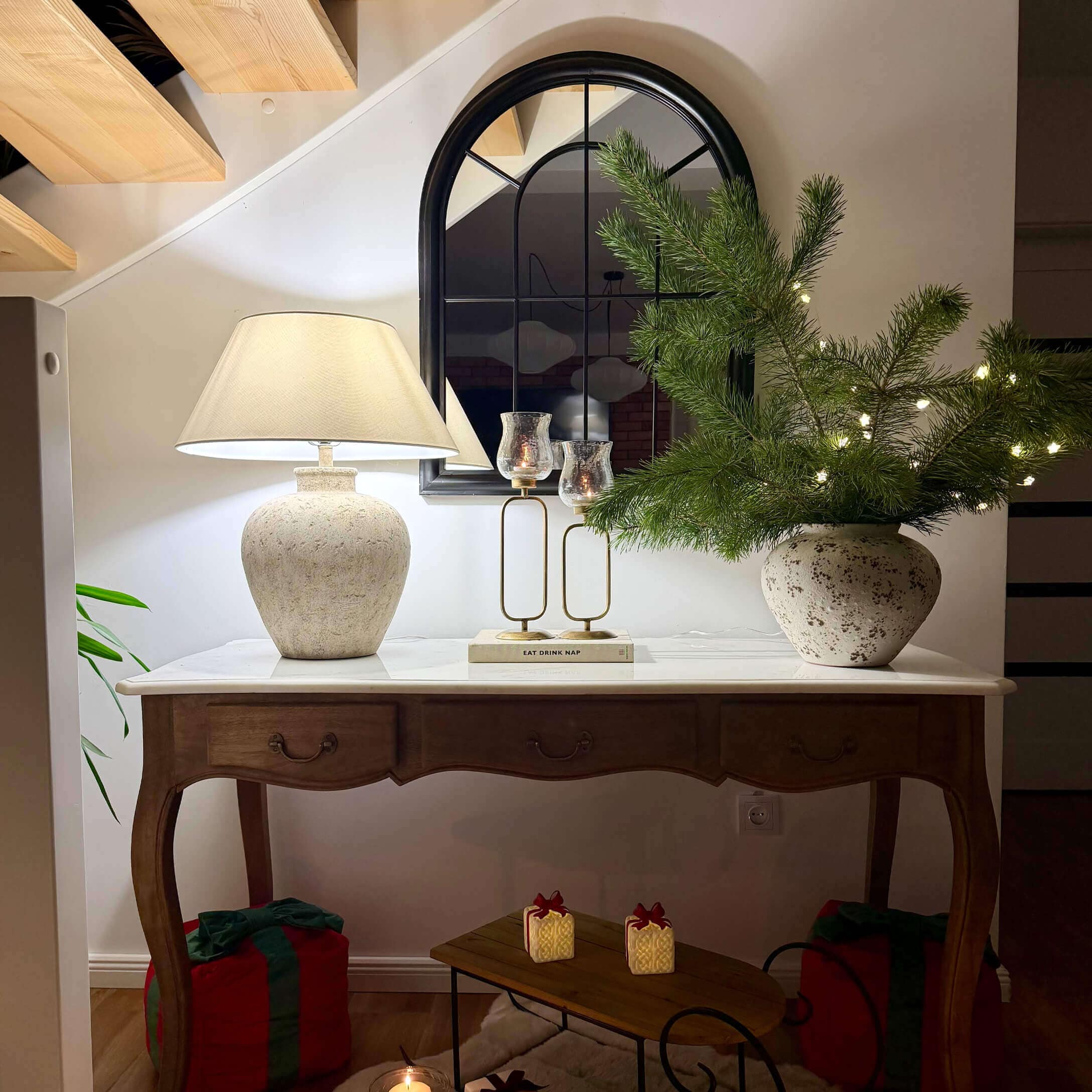 Houten tafel met wit blad, keramische lamp, spiegel en kerstdecoratie met groene takken en lichtjes