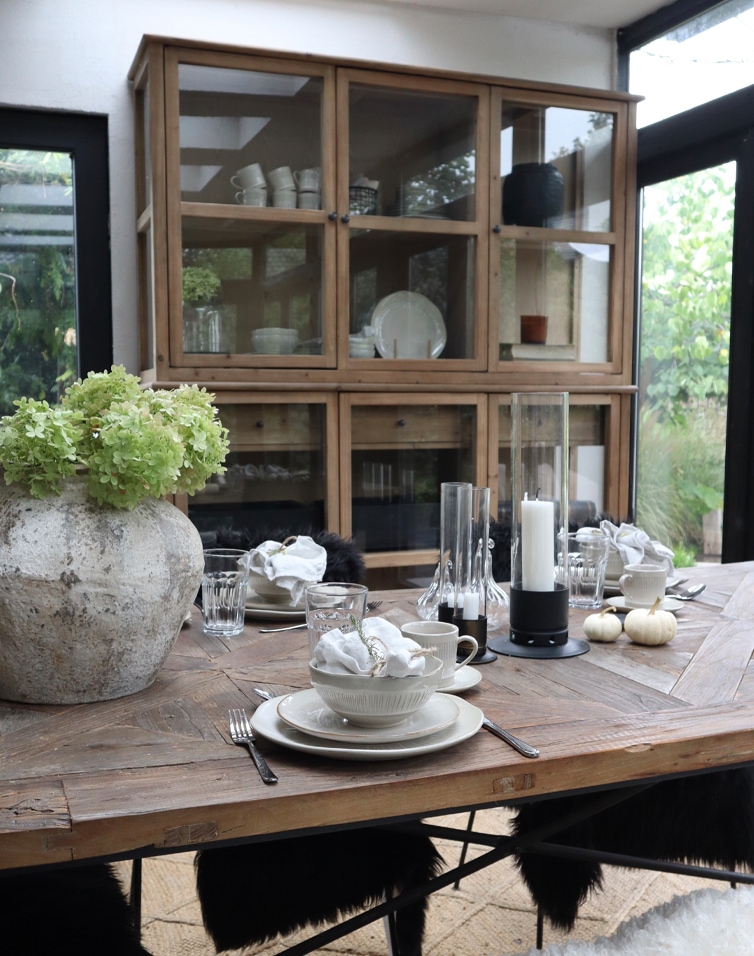 Rustieke houten tafel met servies en grote vaas met groene bloemen in lichte orangerie