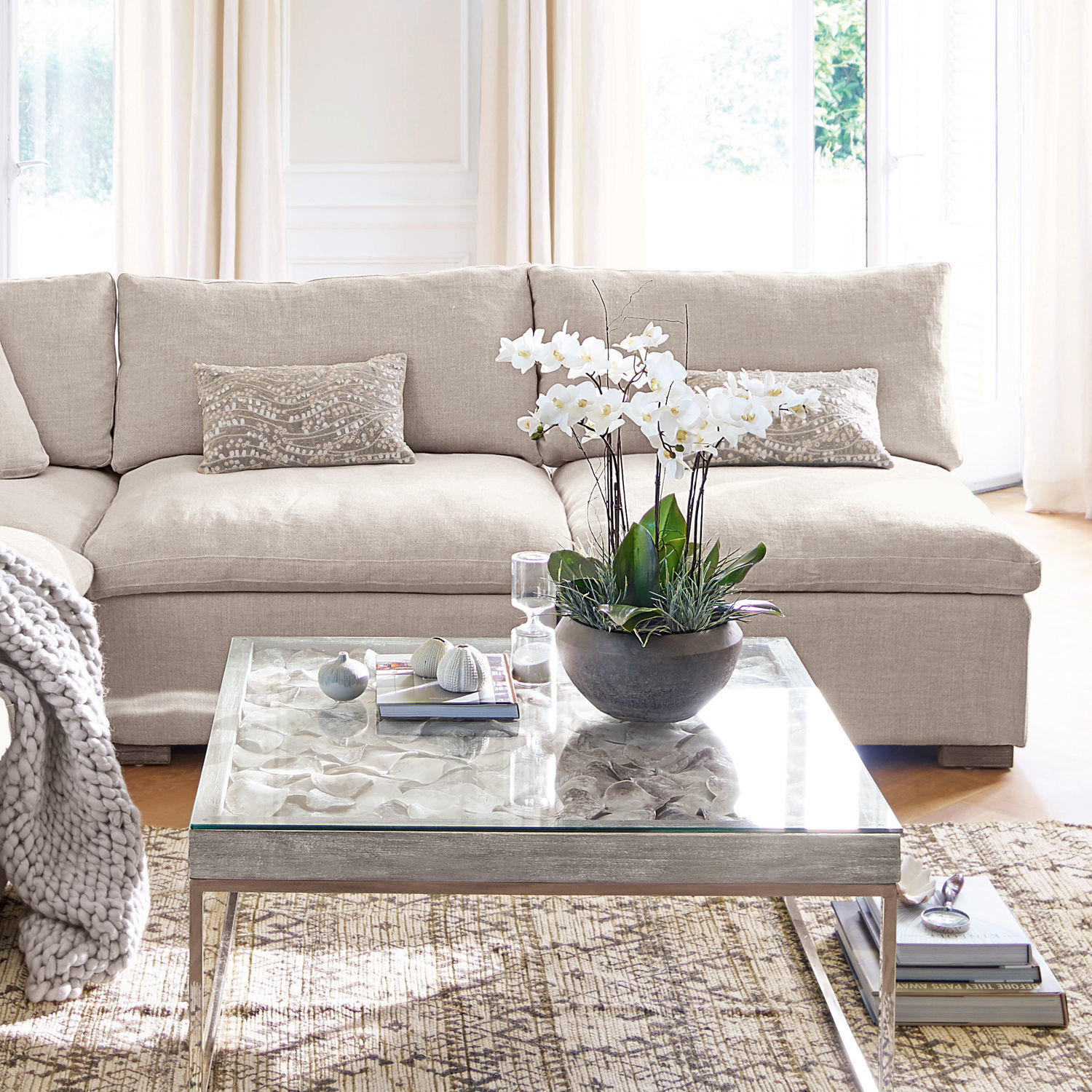 Beige stoffen bank met sierkussens en glazen salontafel met witte bloemen in woonkamer