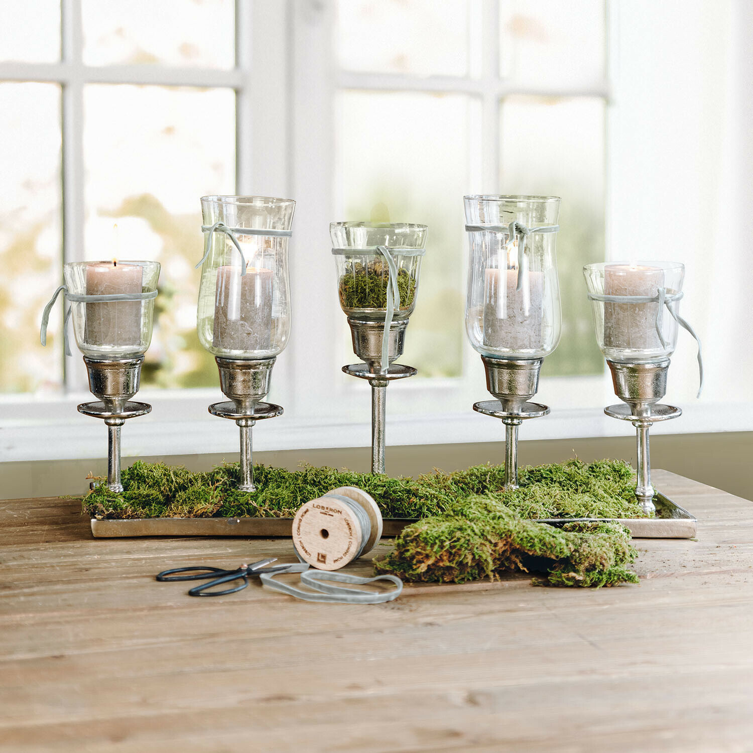 Set van vijf glazen windlichten op metalen standaards met groen mos op een houten tafel