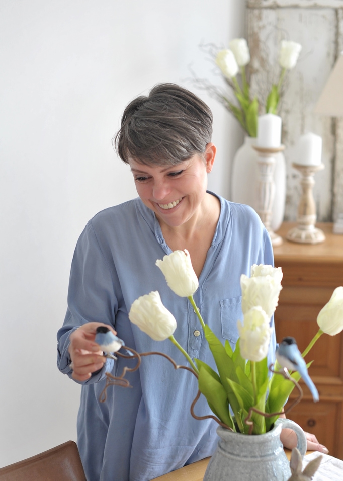 Vrouw in blauw overhemd met witte tulpen en decoratieve vogels in een grijze kan op tafel