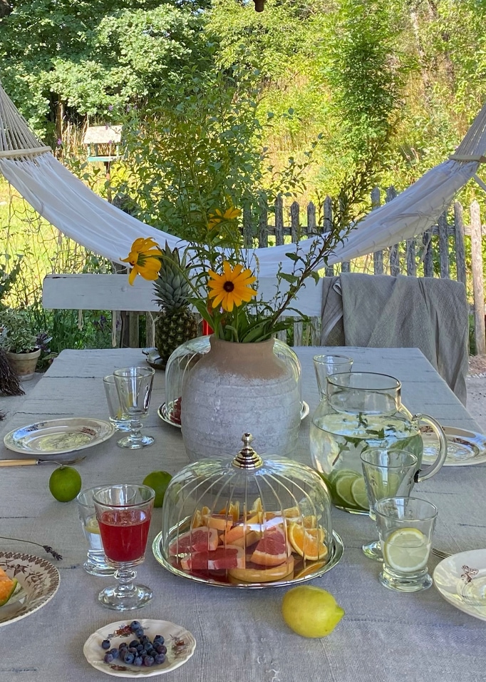 Buitentafel met grijze tafelkleed, vaas met gele bloemen en glazen stolp met fruit, zomerse picknick