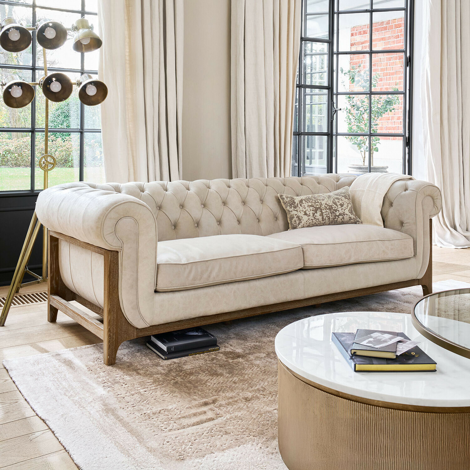 Beige gestoffeerde Chesterfield bank met houten frame in lichte woonkamer met grote ramen en staande lamp
