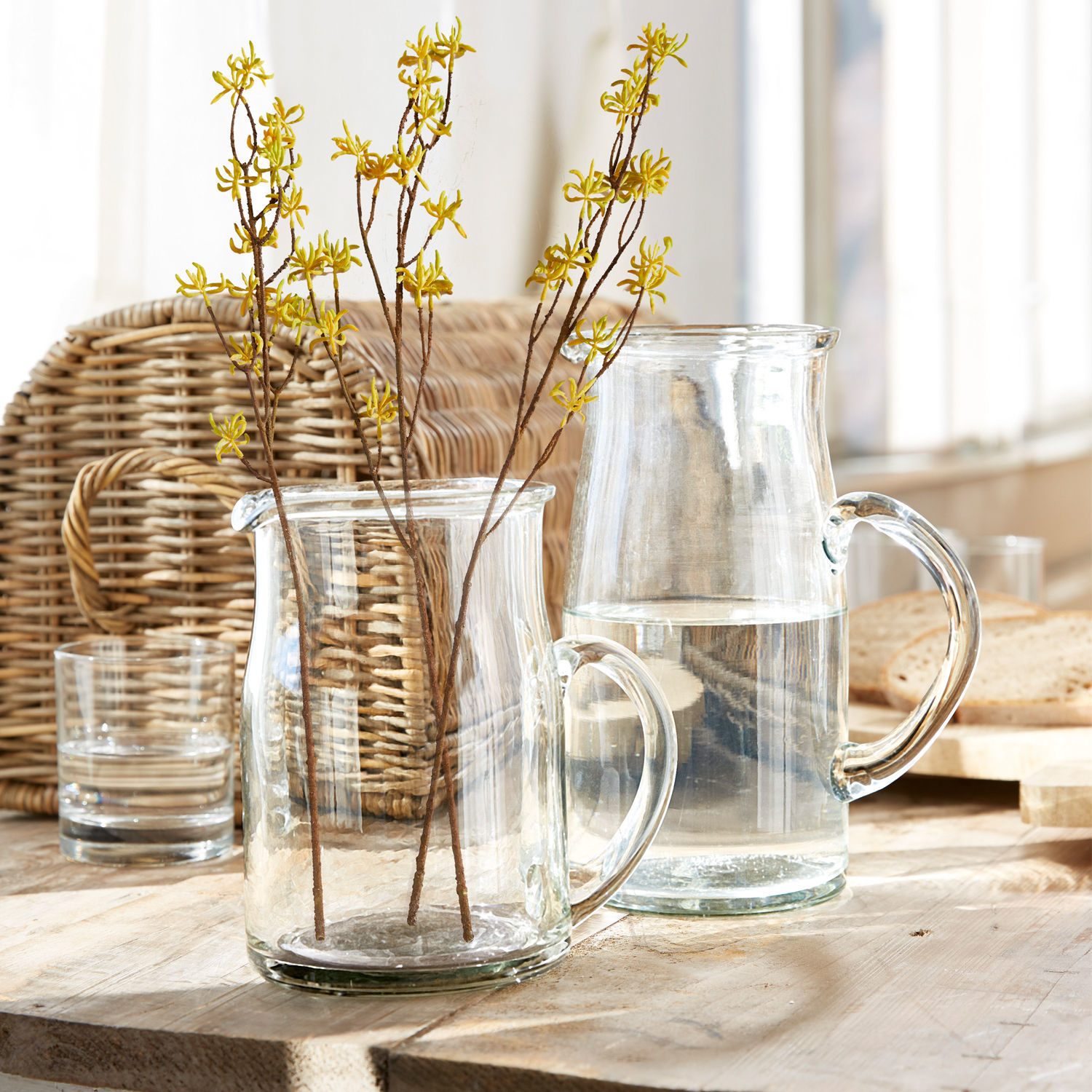 Transparante glazen kan en kan met takken gele bloemen op lichte houten tafel