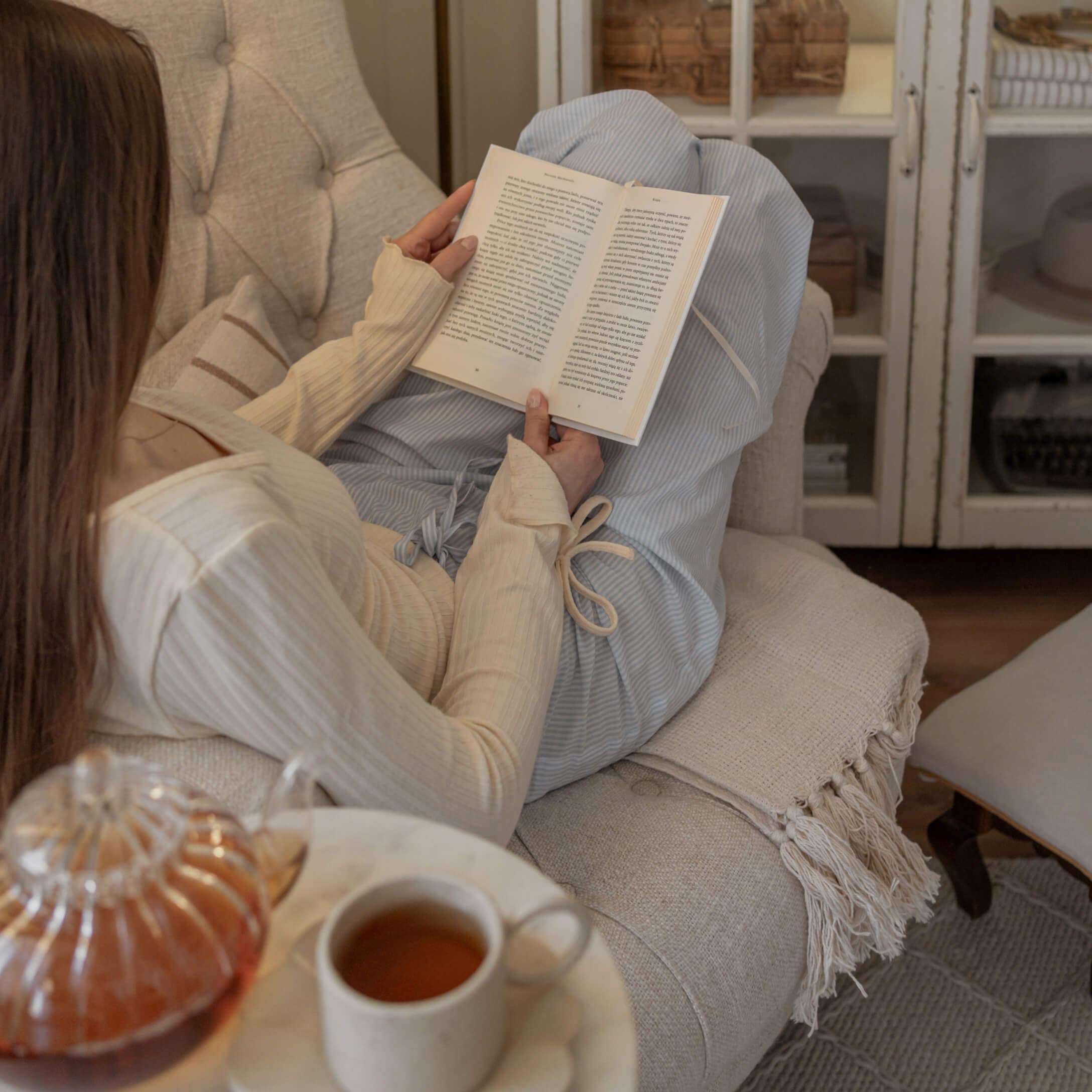 Vrouw in crème trui en blauwe broek leest boek op beige bank met thee op tafel