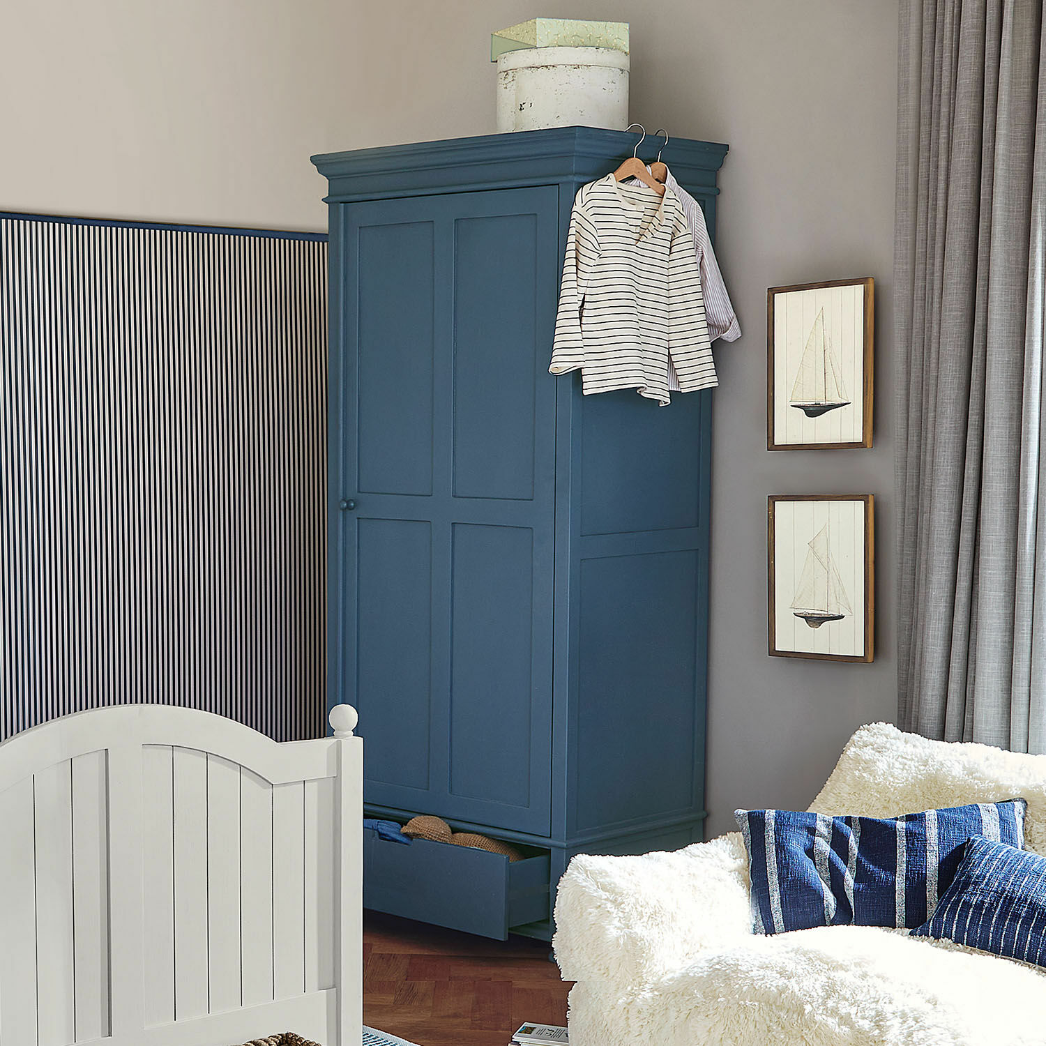 Kledingkast Westcombe Donkerblauwe houten kast met open lade en hangende kleding in gezellige woonkamer met witte bank
