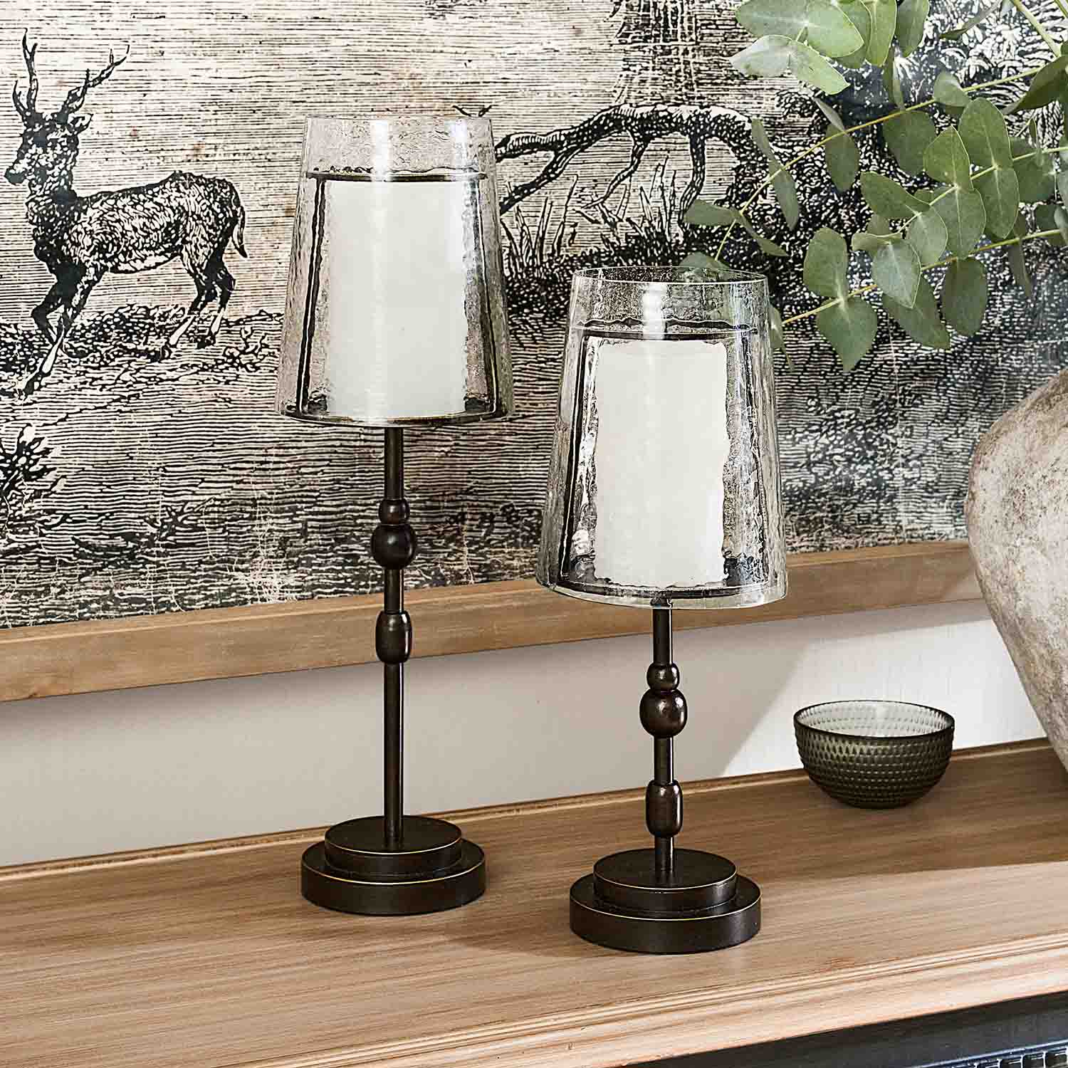 Twee metalen kandelaars met transparante glazen kappen en witte kaarsen op een houten blad