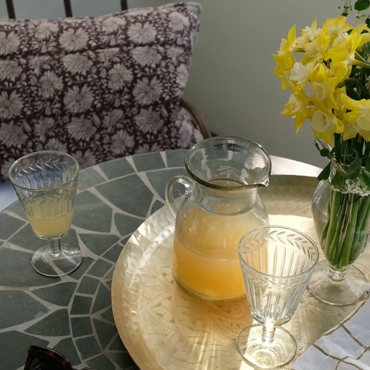 Glazen karaf met gele drank en twee glazen op marmeren tafel met bloemen in vaas
