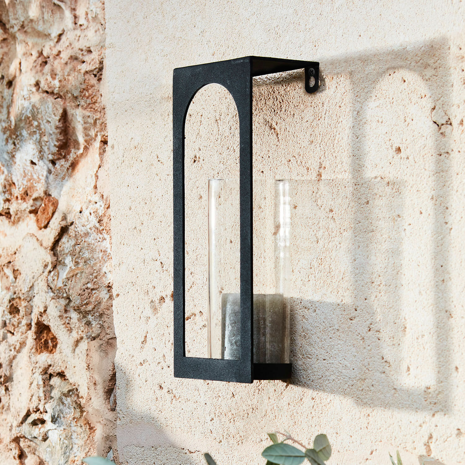 Zwarte metalen wandlamp met doorzichtige glazen houder en kaars aan beige stenen muur