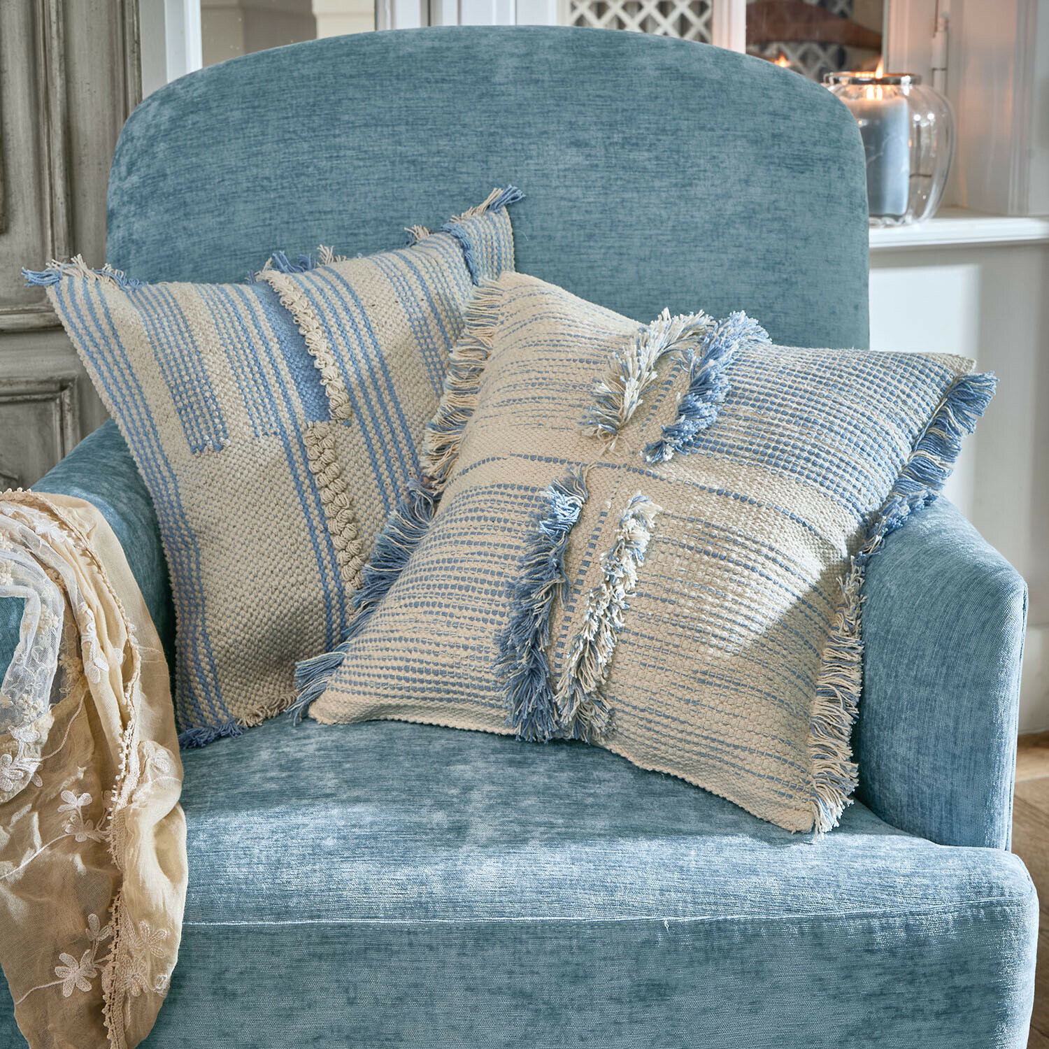 Twee decoratieve katoenen kussens in crème en blauw op een zachte blauwe fauteuil in lichte kamer