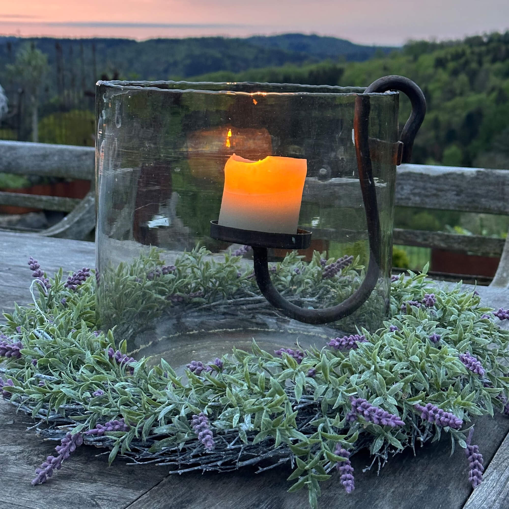 Glazen windlicht met metalen houder en groene krans met paarse bloemen op houten tafel