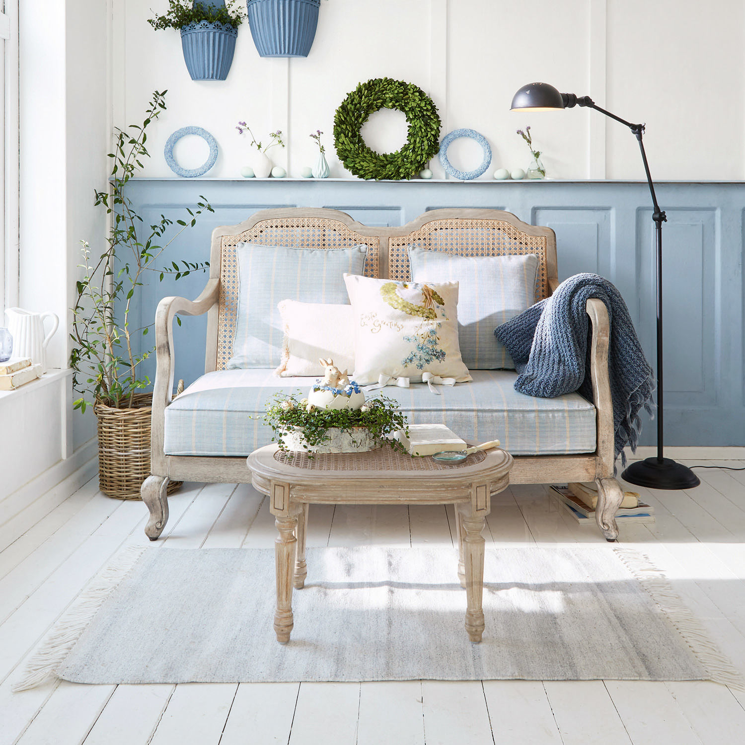 Bank Ainsley Houten rotan bank met blauwe bekleding en kussens in lichte woonkamer met planten en vloerlamp