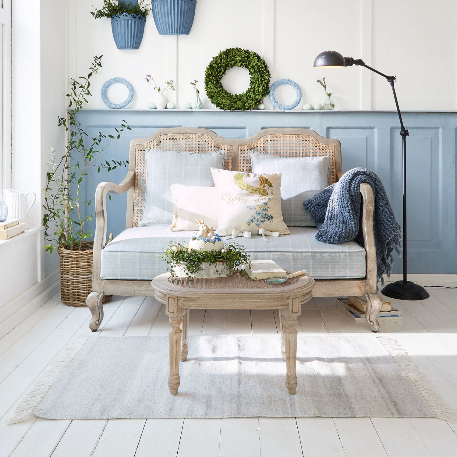 Bank Ainsley Houten rotan bank met blauwe bekleding en kussens in lichte woonkamer met planten en vloerlamp