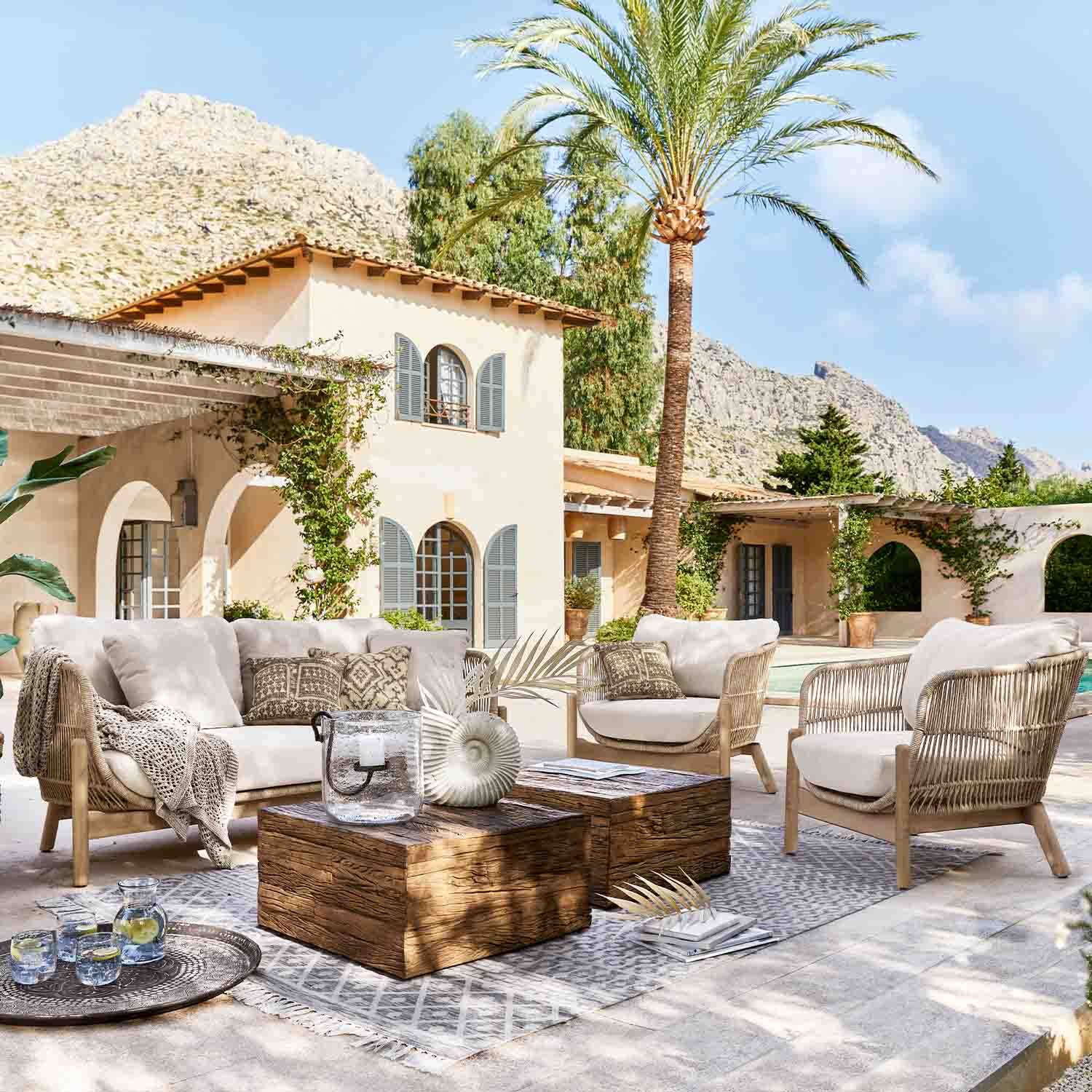 Buitenset van licht rattan met beige kussens op terras met palmboom en bergzicht